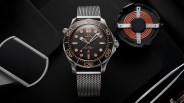 Omega Seamaster wristwatch with a black dial, beige hour markers, and a stainless steel mesh strap, displayed on a black surface alongside a copper coil component.
