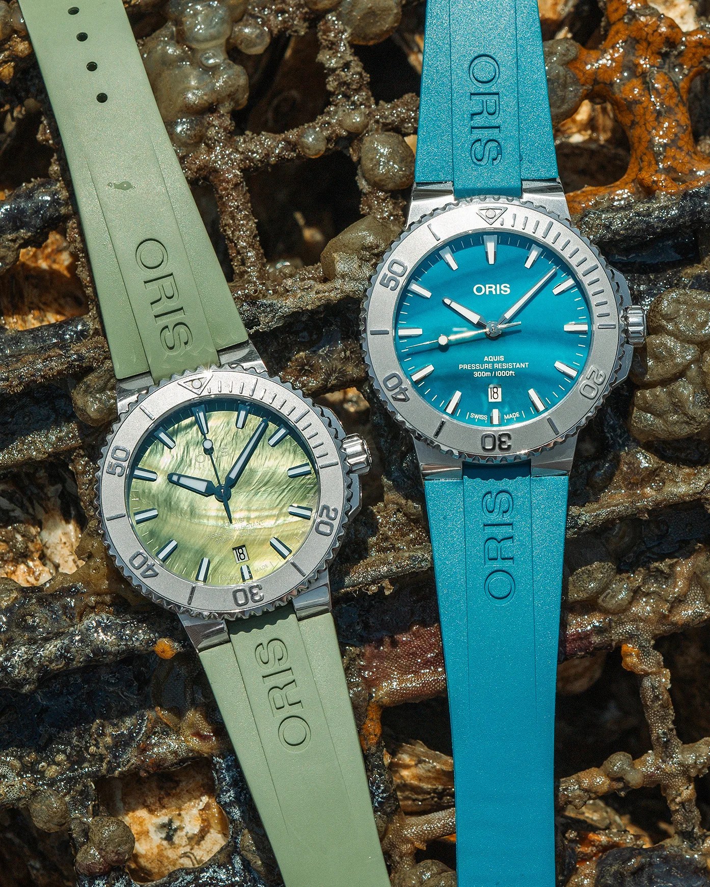Two Oris wristwatches with rubber straps are displayed on a textured, natural background. The watch on the left has a light green strap and a matching green mother-of-pearl dial with silver hour markers and hands. The watch on the right features a bright blue strap and a blue dial with silver hour markers and hands.