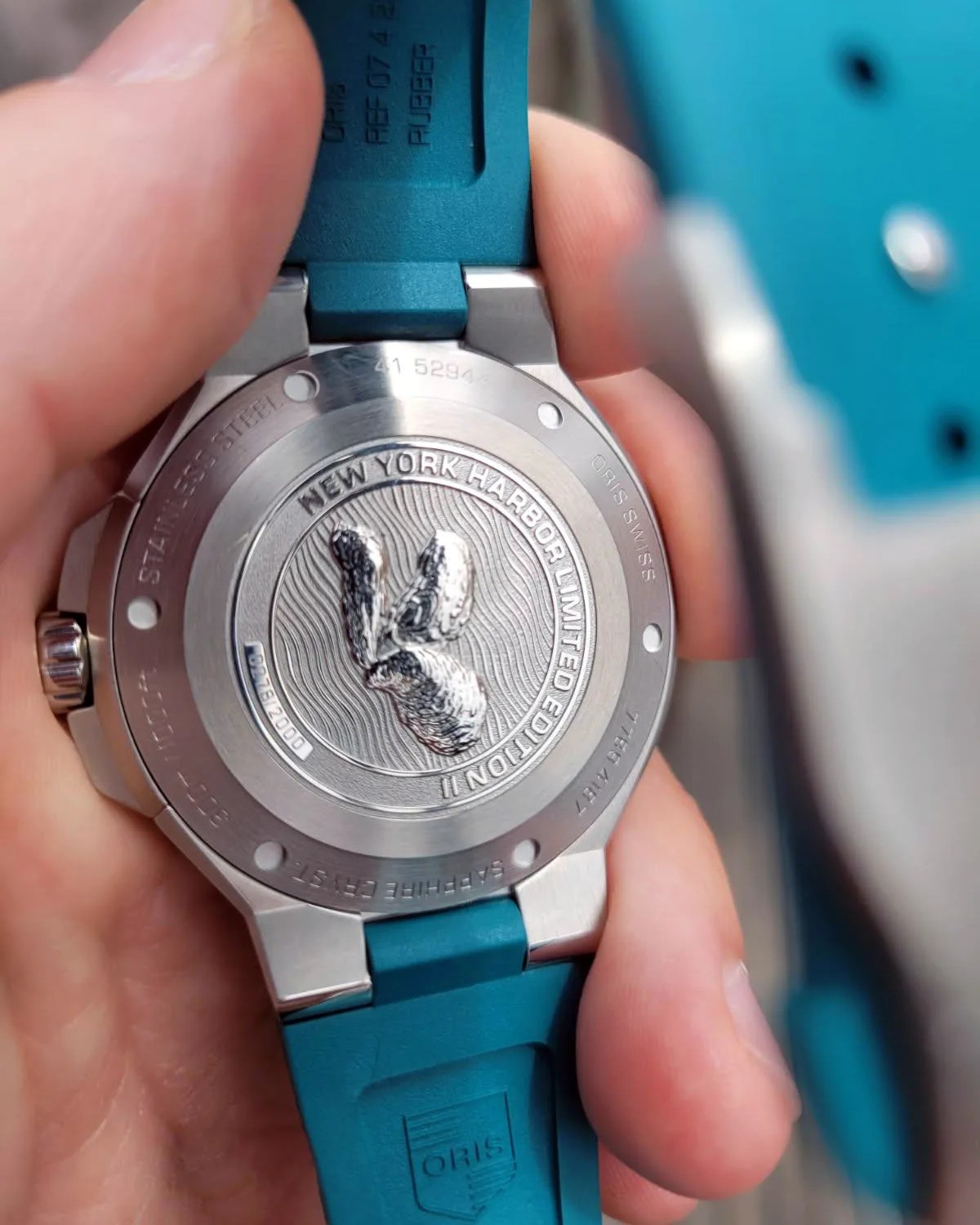 Back of a stainless steel wristwatch with a teal rubber strap, featuring an engraved emblem and text that reads "NEW YORK HARBOR LIMITED EDITION II" and "00278/2000".