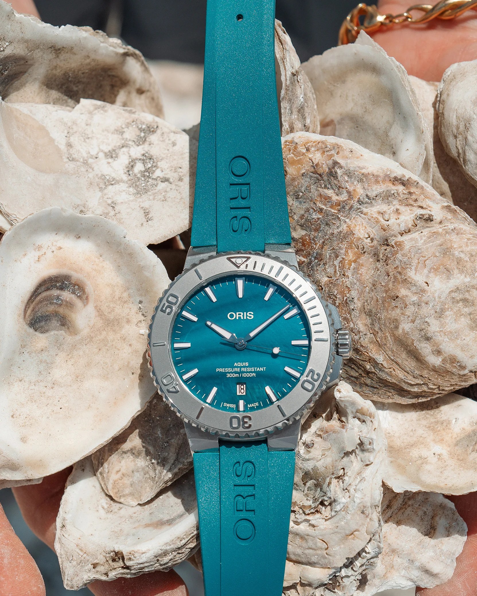 Blue Oris Aquis dive watch with a stainless steel bezel and blue rubber strap, placed on a pile of oyster shells.