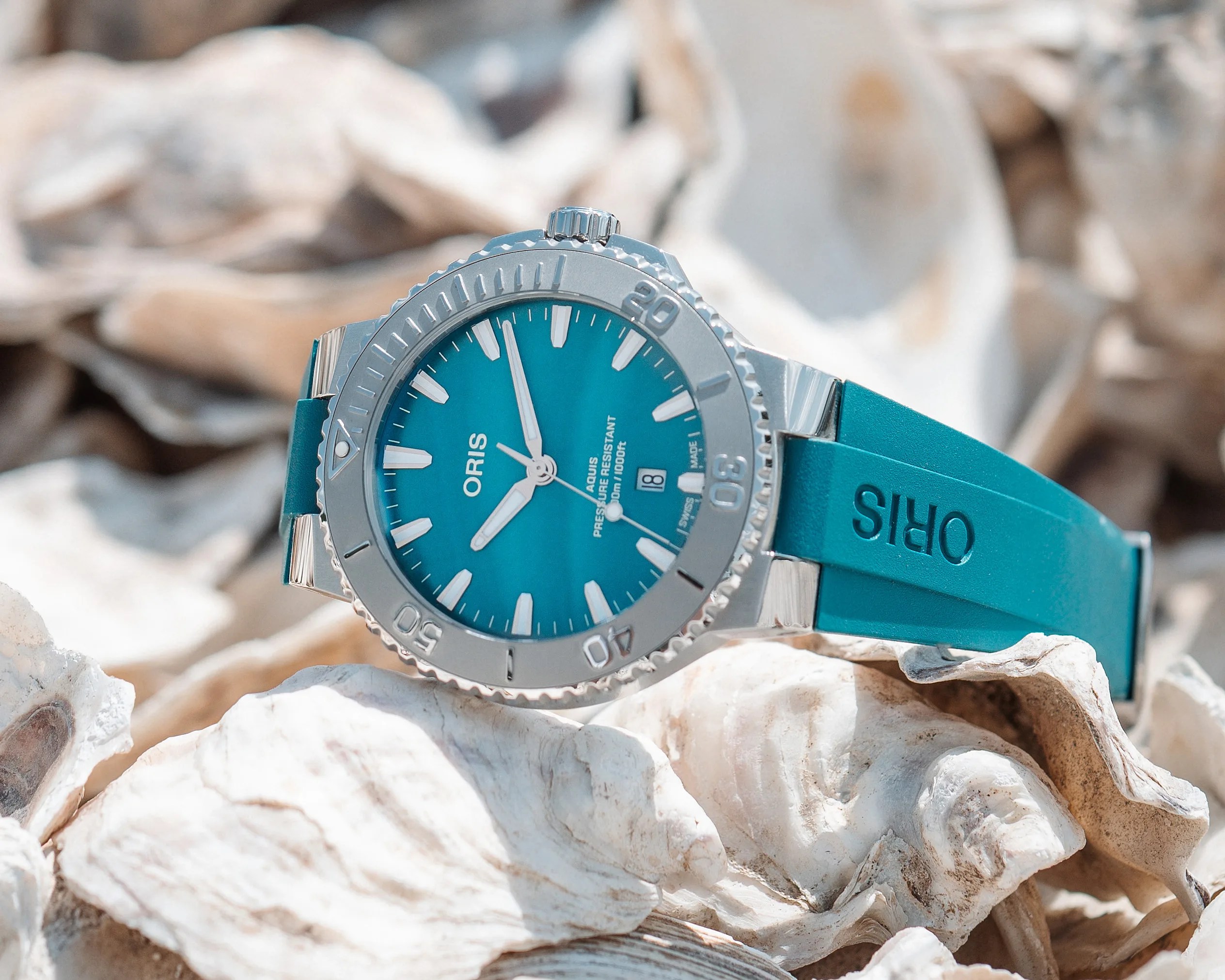 A steel Oris wristwatch with a turquoise dial and matching turquoise rubber strap, placed on a bed of white seashells. The watch face features white hour markers, a date window at 6 o'clock, and the text "ORIS," "AQUIS," and "PRESSURE RESISTANT 30 bar / 300 m / 1000 ft."