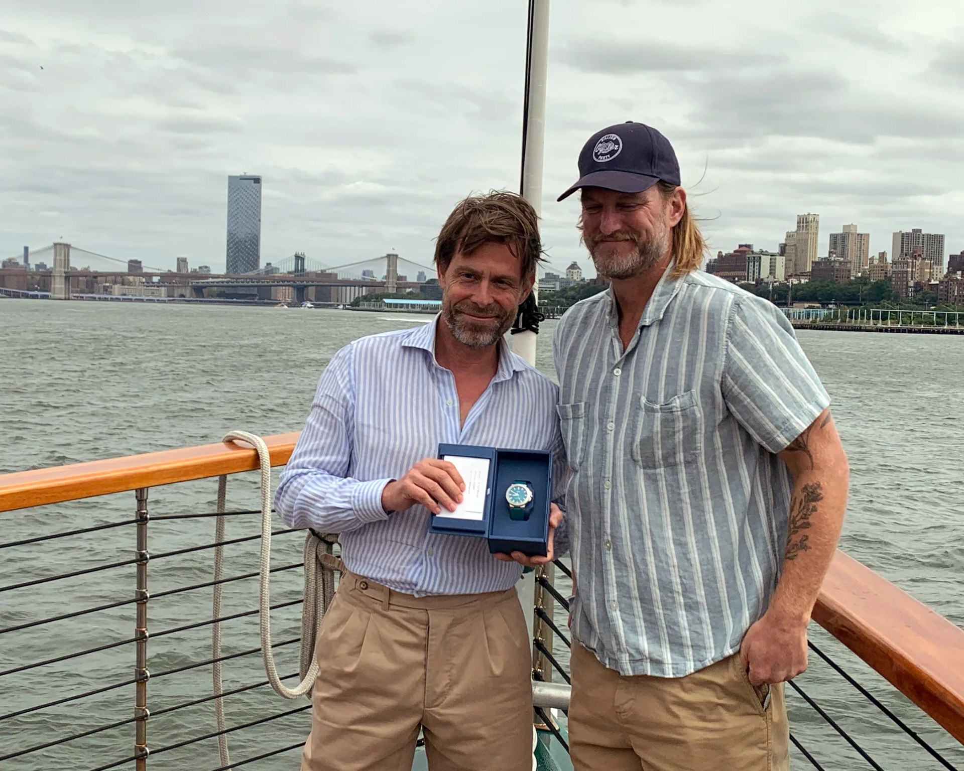Two men are standing on a boat with the Brooklyn Bridge and the Williamsburg Bridge behind them.
