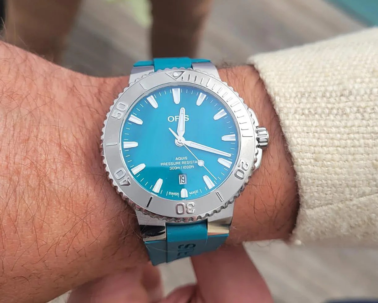 Blue-dial Oris Aquis wristwatch with a silver bezel and blue rubber strap, displaying the time as 12:15:30, worn on a wrist with a beige textured sleeve. The watch face includes white hour markers, a date window at 6 o'clock, and text indicating "Aquis Pressure Resist 300m/1000ft" and "Swiss Made.