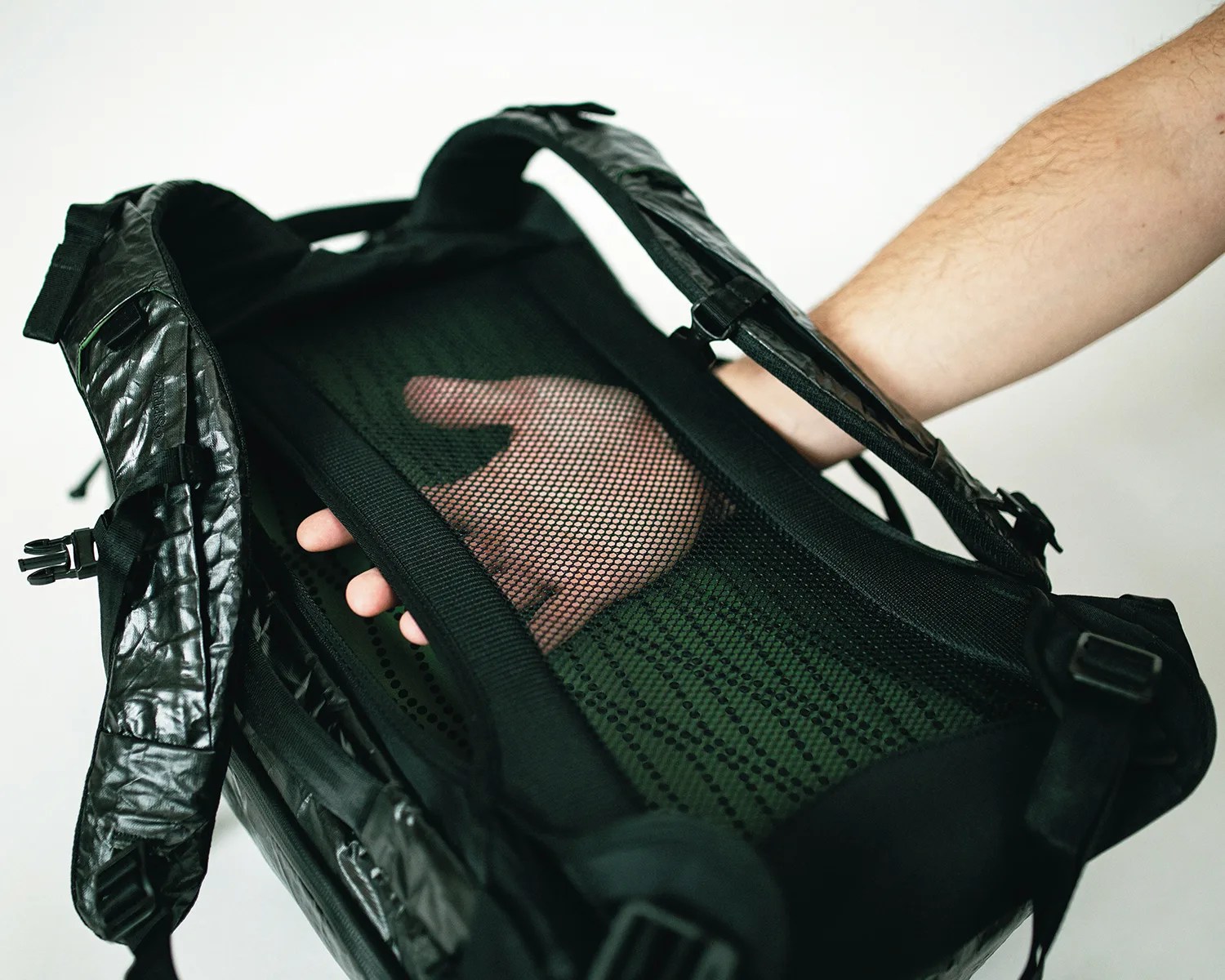 Black backpack with padded shoulder straps and a mesh back panel, showing a hand placed behind the mesh for ventilation demonstration. The backpack has adjustable straps and buckles.