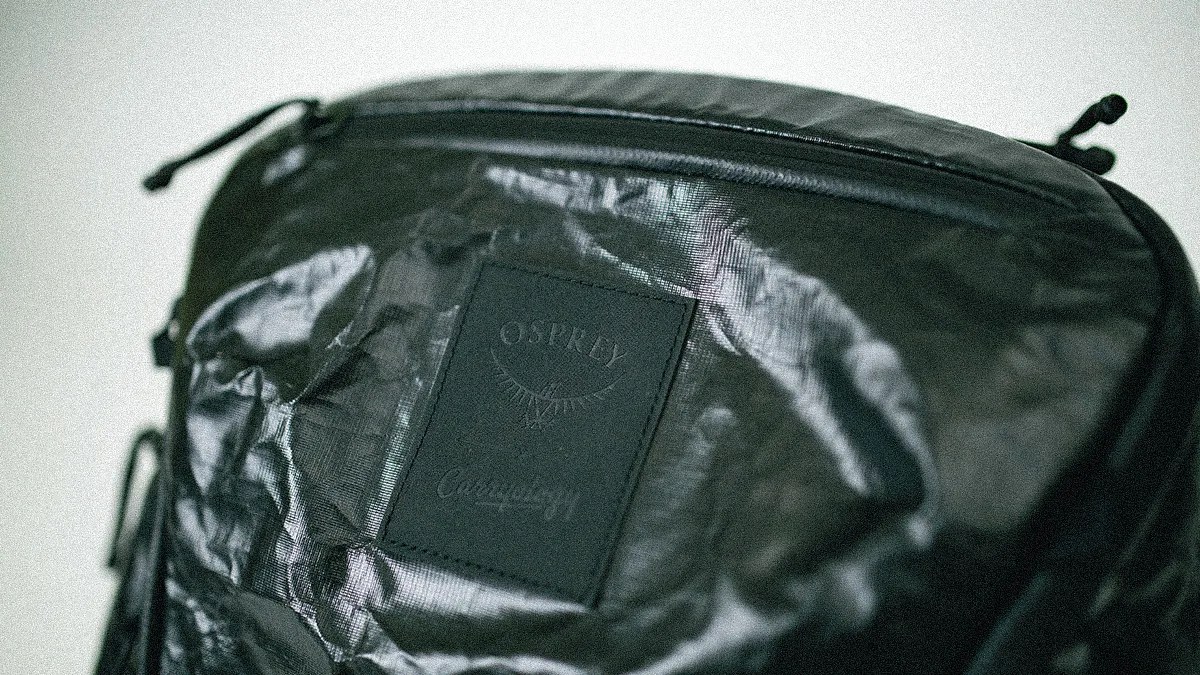Black Osprey backpack with a textured, slightly shiny fabric and a stitched rectangular patch featuring the Osprey logo and text.