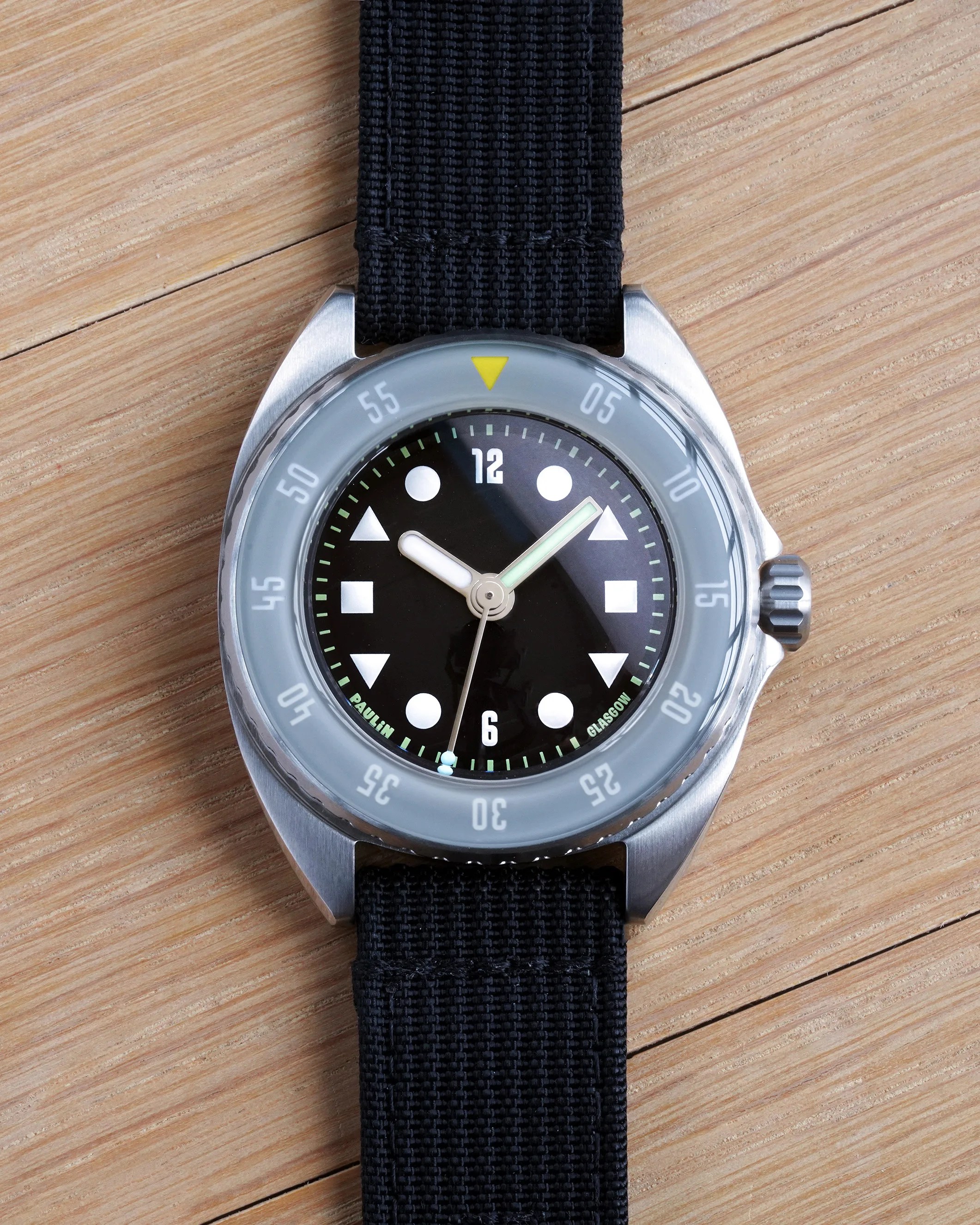 A wristwatch with a black dial featuring white geometric hour markers (circles, triangles, and squares) and white numerals at 12 and 6. The watch has a silver stainless steel case, a gray bezel with white minute markings, and a yellow triangle at the 12 o'clock position. The hour and minute hands are white with luminescent centers, and the second hand is thin and silver. The watch is attached to a black woven fabric strap and is placed on a light wood surface. The words "PAULIN" and "GLASGOW" are printed in green near the 7 and 5 o'clock positions on the dial.