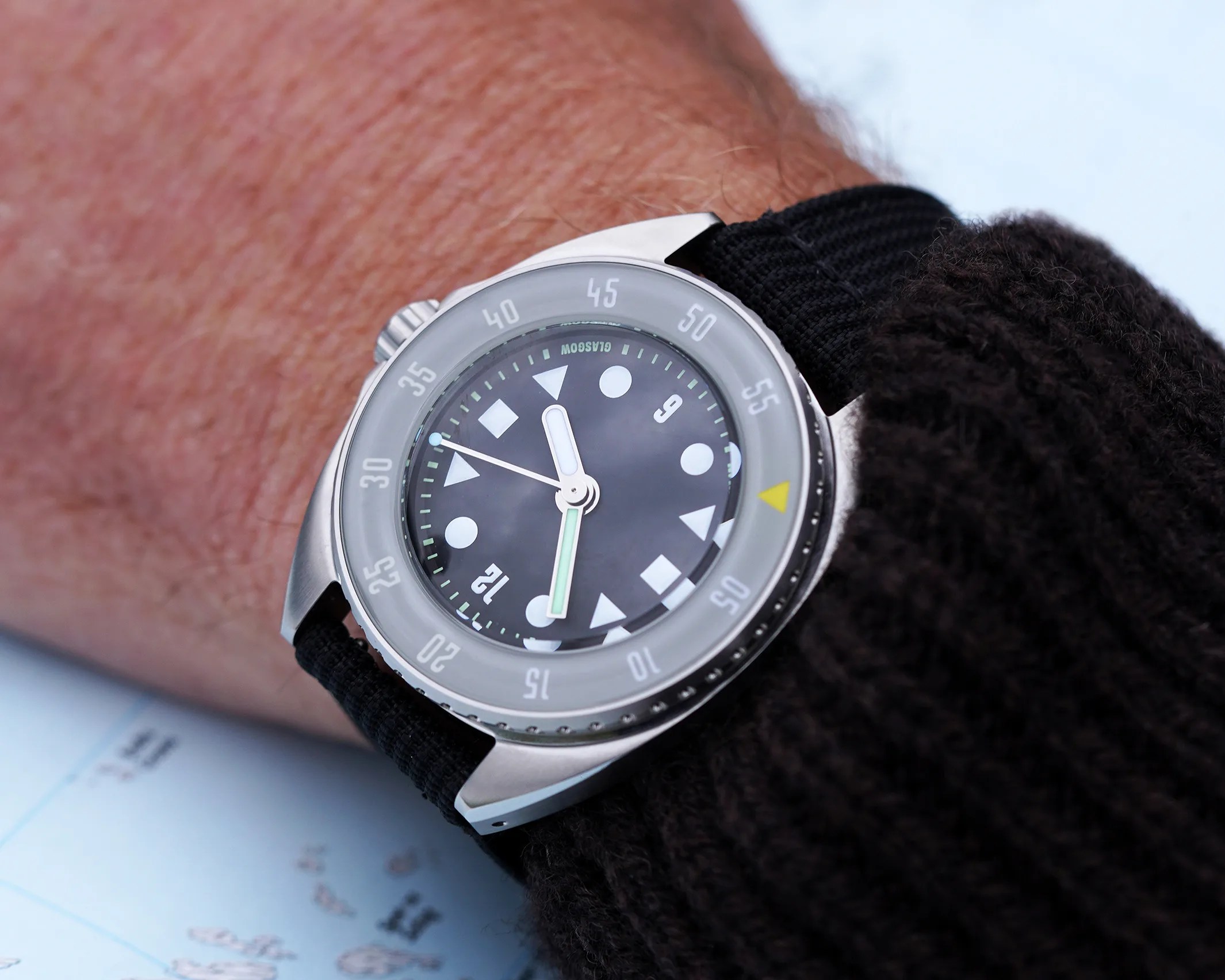 A wristwatch with a black dial featuring geometric hour markers (circles, triangles, and diamonds) and a black fabric strap. The watch has a silver bezel with minute markings and a yellow triangle at the 12 o'clock position. The watch is worn on a wrist with a dark knitted sleeve visible.