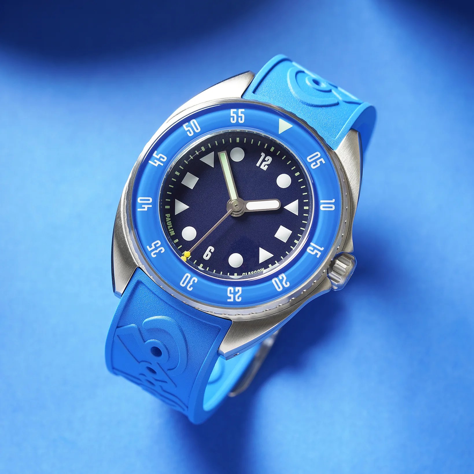 Blue wristwatch with a stainless steel case, blue bezel marked with white numbers, and a blue rubber strap embossed with a design. The watch face is dark blue with white geometric hour markers and white hour and minute hands. The second hand is thin and silver with a small yellow detail near the tip.