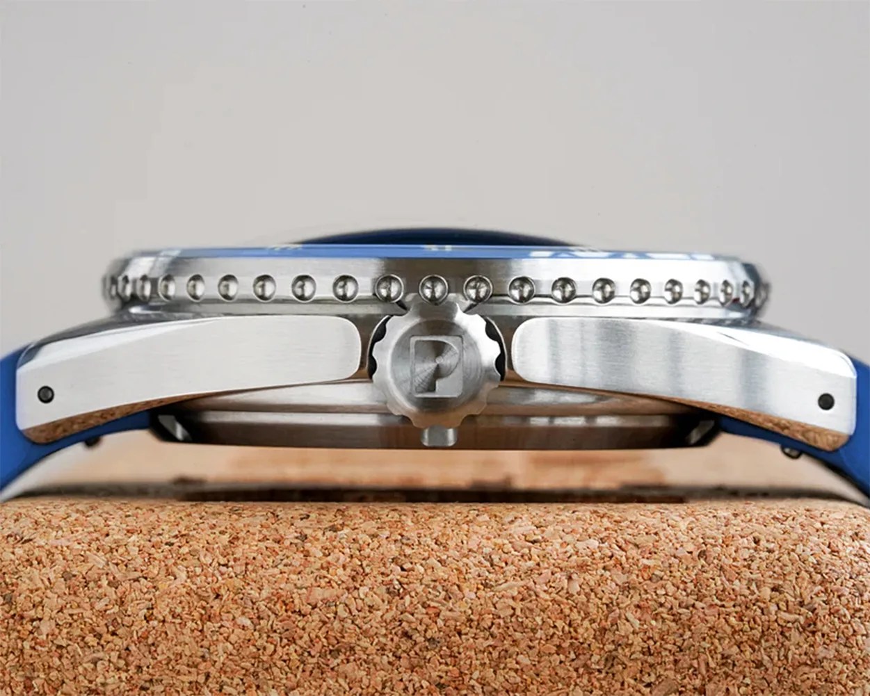 Side view of a stainless steel watch case with a textured bezel featuring evenly spaced round studs. The crown has a "P" logo engraved on it. The watch is mounted on a blue strap and rests on a cork surface.