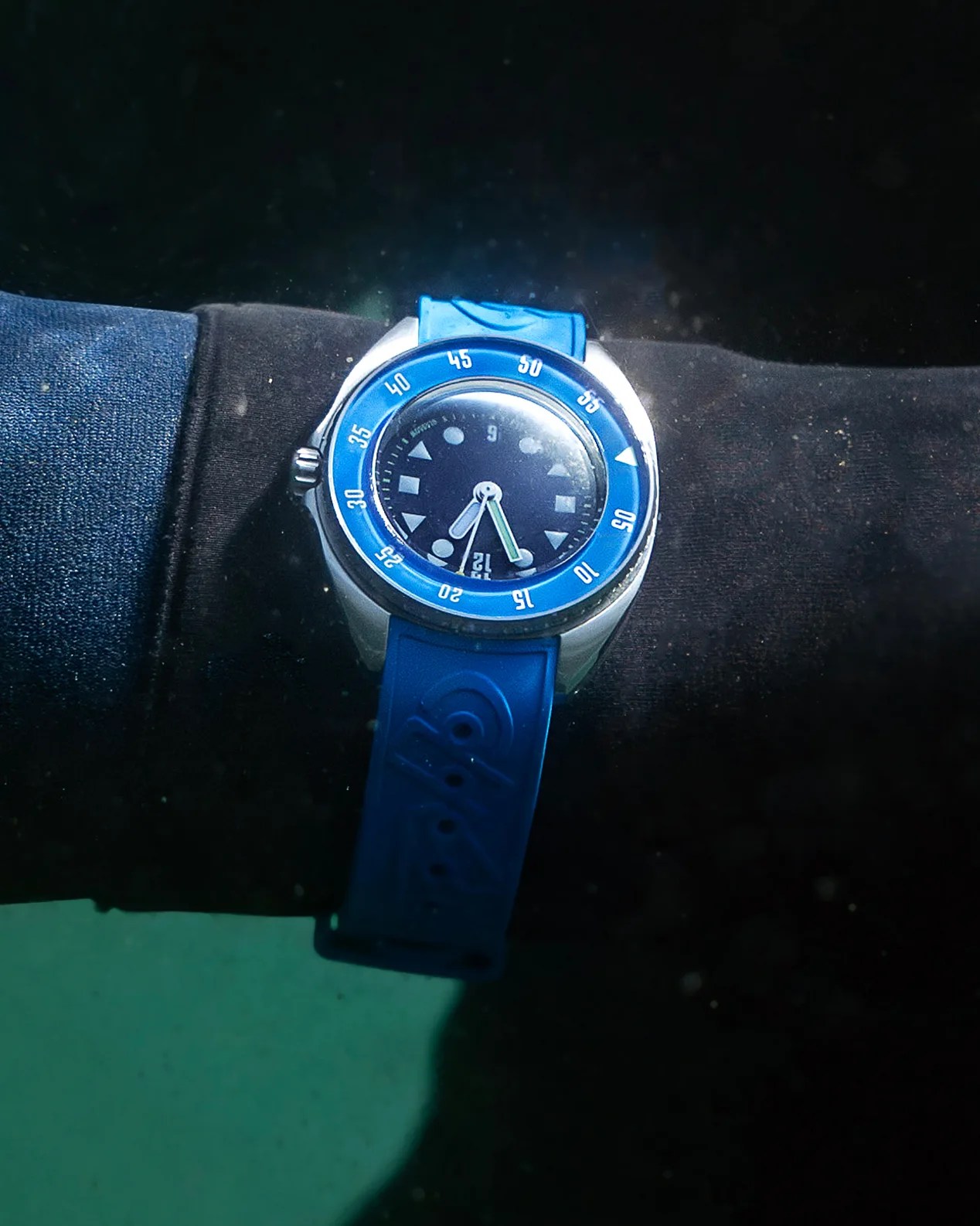 A wristwatch with a blue bezel and blue rubber strap is worn over a black wetsuit sleeve. The watch face is dark with white geometric hour markers and white hands, and the bezel has white minute markings. The watch appears to be underwater.