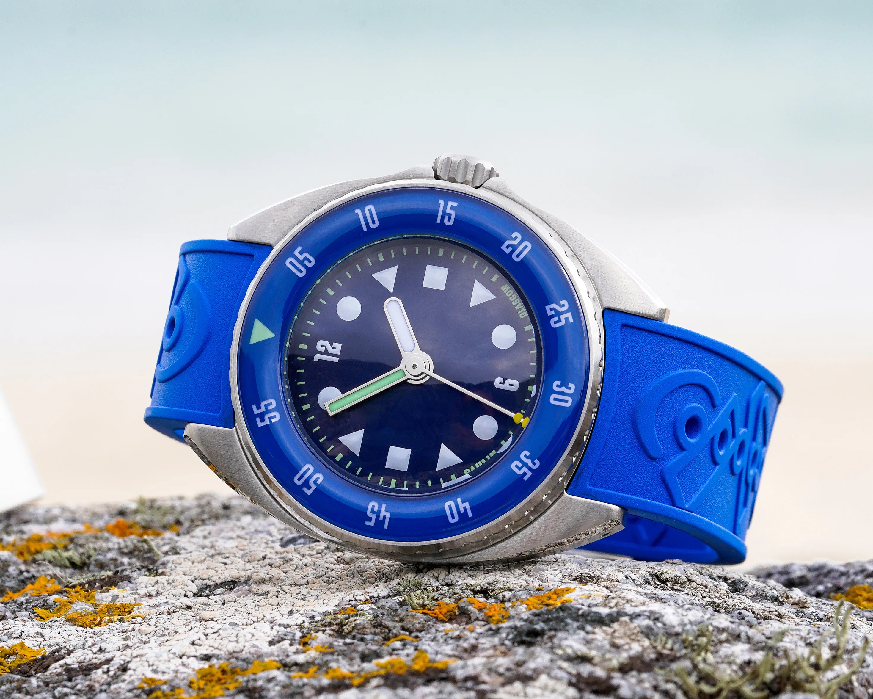 Blue wristwatch with a stainless steel case, featuring a blue bezel marked with white numerals in increments of five. The watch face is dark blue with white geometric hour markers and luminous hands. The strap is blue rubber with embossed designs. The watch is resting on a textured rock surface with some orange and green lichen.