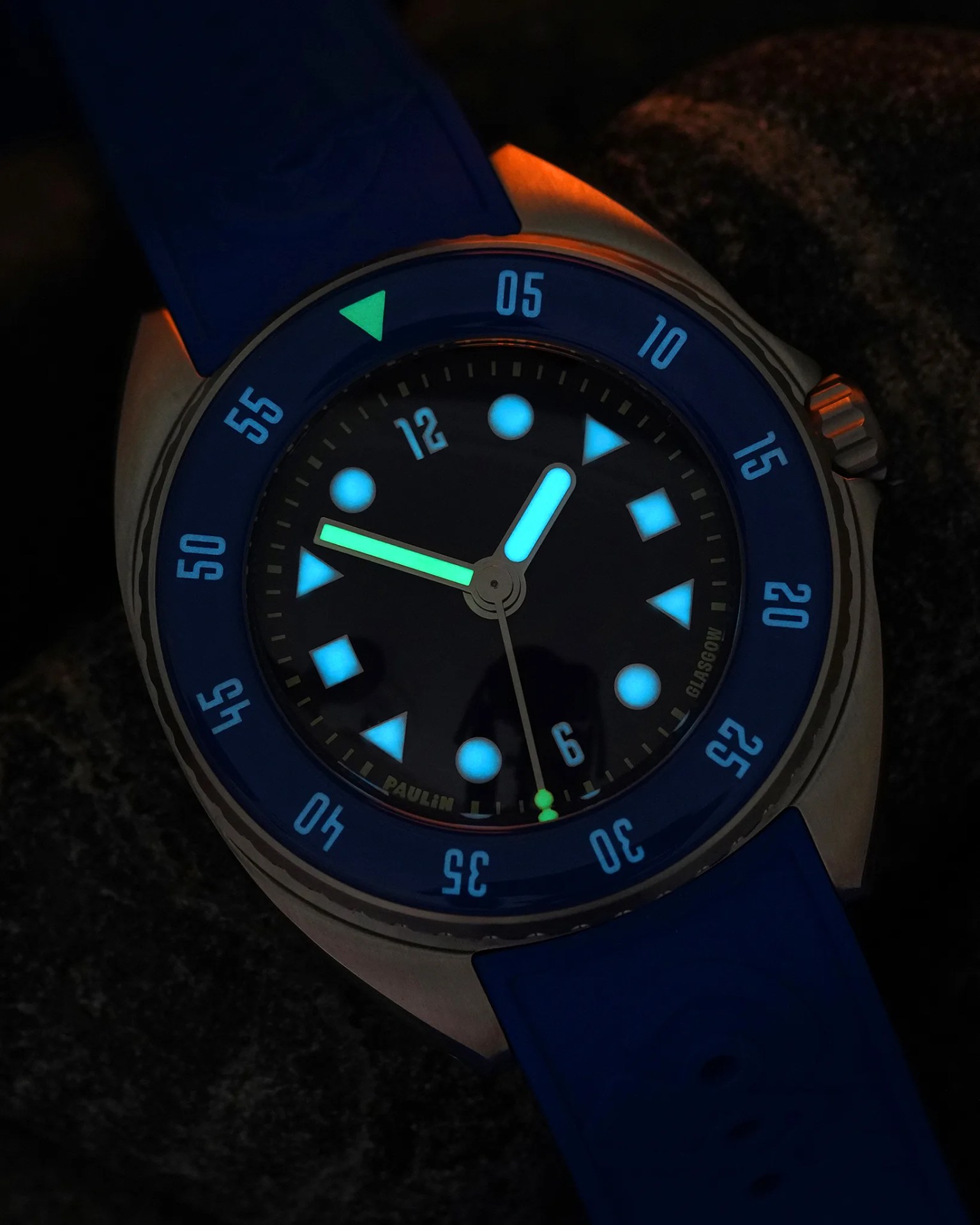 Diver-style wristwatch with a blue bezel and blue rubber strap. The watch face is black with luminescent hour markers in various shapes (circles, triangles, and diamonds) glowing blue. The hour hand glows blue, and the minute hand glows green. The bezel has minute markers in light blue, and the watch case is metallic. The brand name "PAULIN" and "GLASGOW" are subtly visible on the dial. The watch is photographed in low light, highlighting the luminescence.