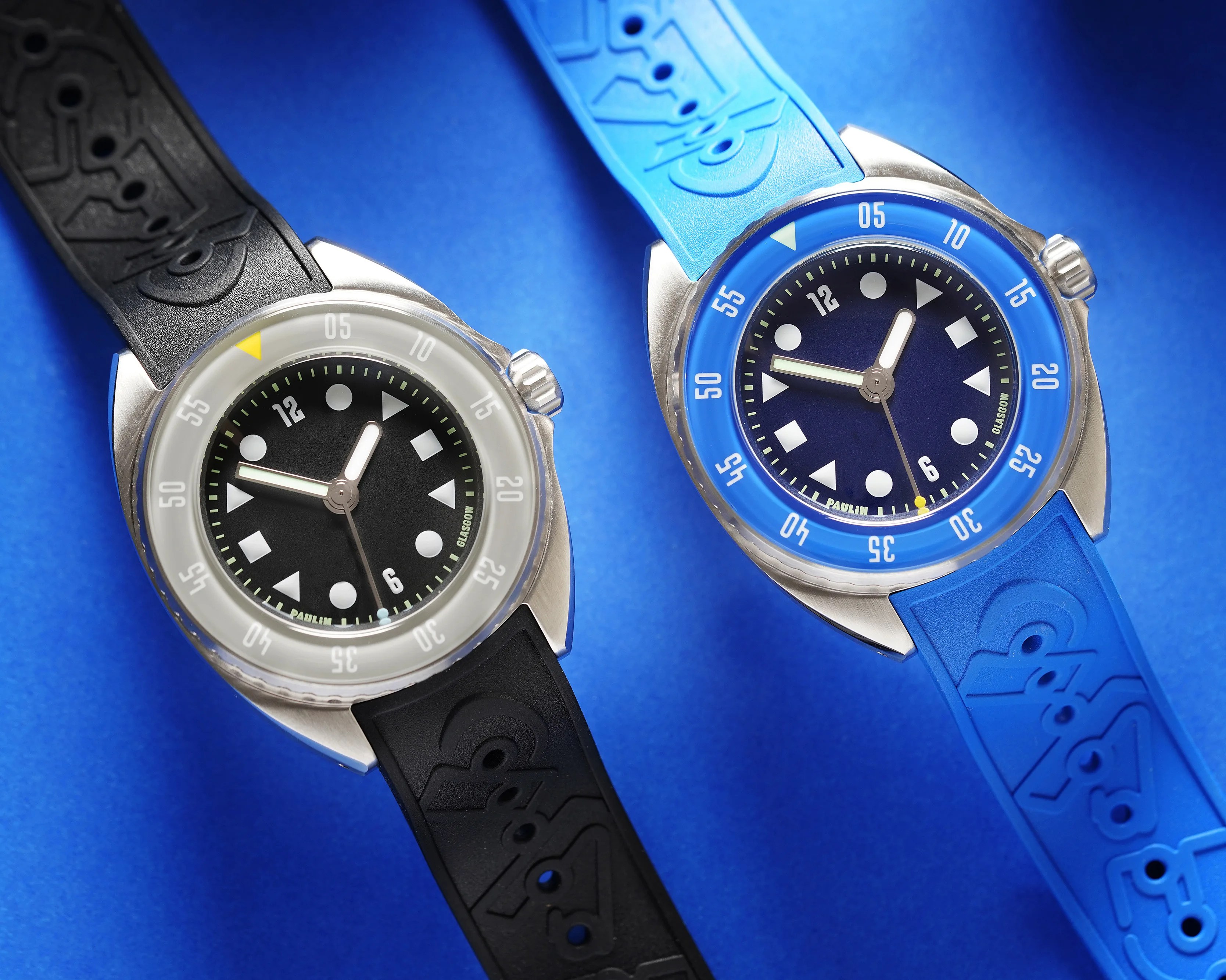 Two wristwatches with round stainless steel cases and rubber straps are shown on a blue background. The watch on the left has a black dial with white geometric hour markers, a translucent bezel with white numerals, and a black strap embossed with a pattern. The watch on the right has a dark blue dial with similar white geometric hour markers, a bright blue bezel with white numerals, and a matching blue strap with an embossed pattern. Both watches have three hands and a crown on the right side.