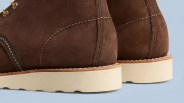 Close-up of the back portion of brown suede boots with white stitching, a light tan welt, and thick off-white rubber soles. The boots have metal eyelets with yellow and red laces visible at the top left. The background is a light blue surface.