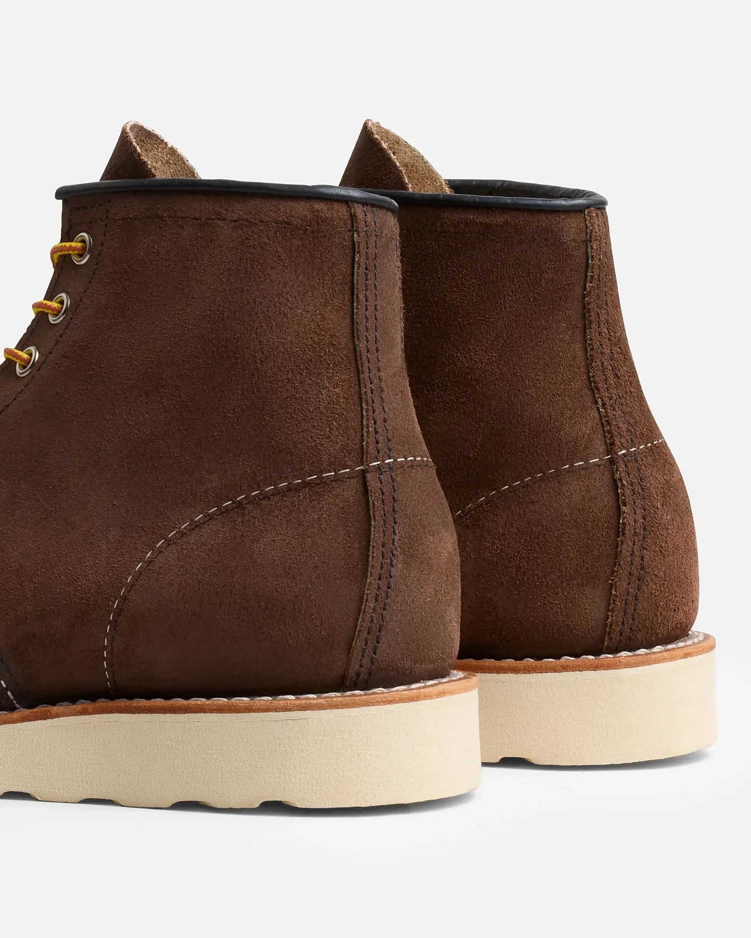 Brown suede boots with white stitching details and cream-colored thick rubber soles. The boots have a black trim around the top edge and yellow and red laces visible on the side. The image shows the back and side view of the boots.