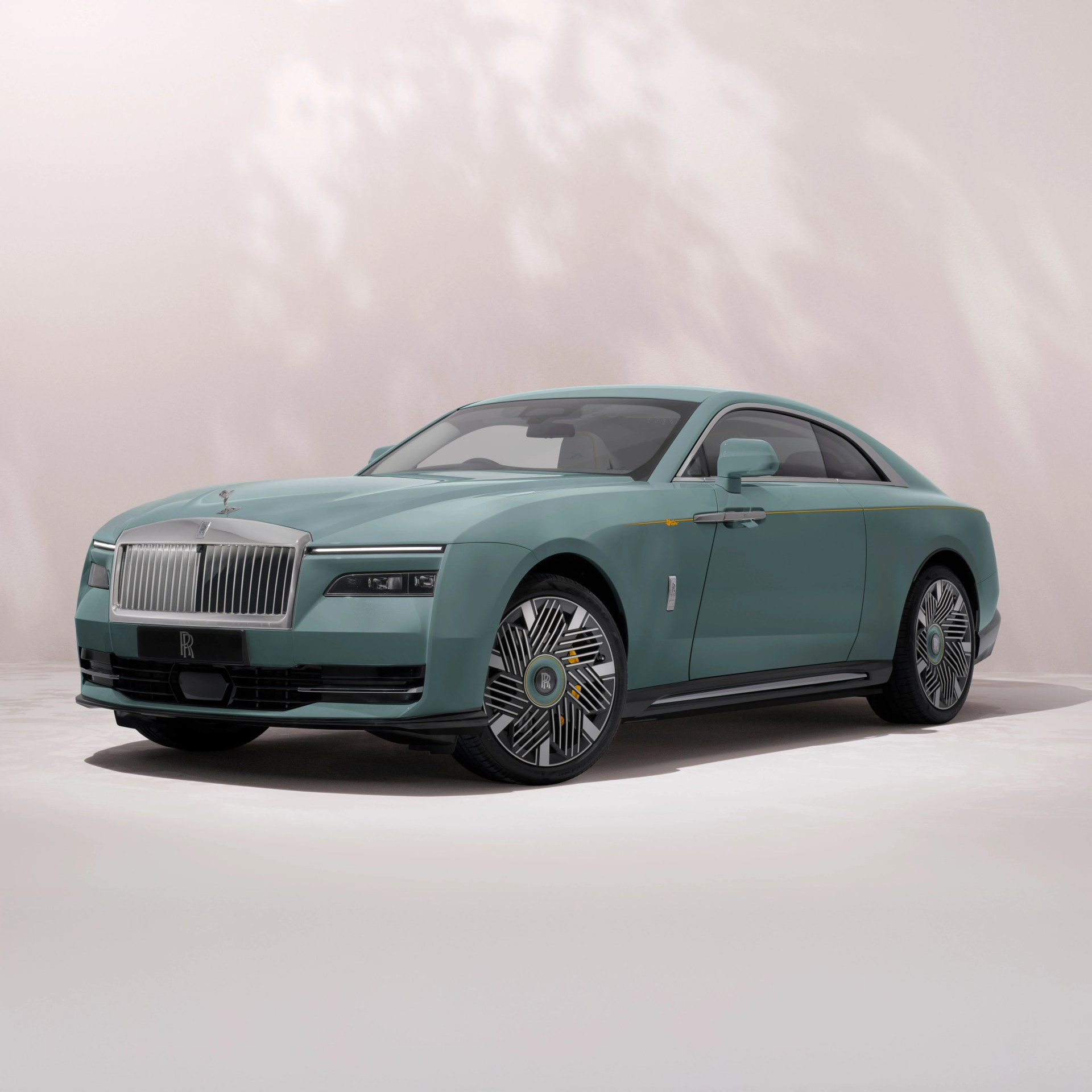 A sleek, modern Rolls-Royce coupe in a muted teal color with a prominent silver grille and the iconic Spirit of Ecstasy hood ornament. The car features distinctive, intricate alloy wheels with the Rolls-Royce logo at the center and a subtle gold pinstripe running along the side. The background is a soft, light neutral tone with gentle shadows.