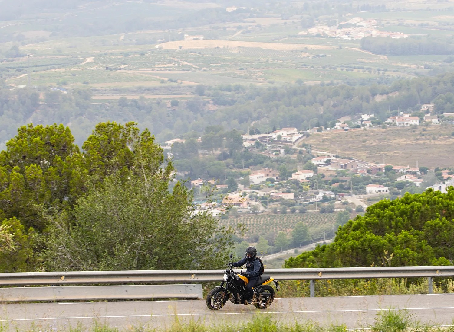 A motorcyclist wearing a black helmet and dark clothing rides a yellow and black motorcycle on a winding road with metal guardrails. The background features a scenic view of a valley with scattered houses, trees, and agricultural fields.