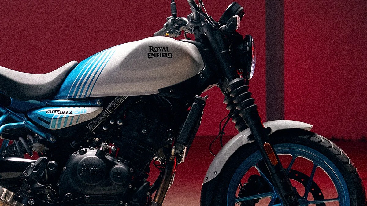 Close-up of a Royal Enfield motorcycle, specifically the Guerilla 350 model, featuring a white fuel tank with blue stripes, a black engine, and a front wheel with a white fender and blue rim. The background is a solid dark red.