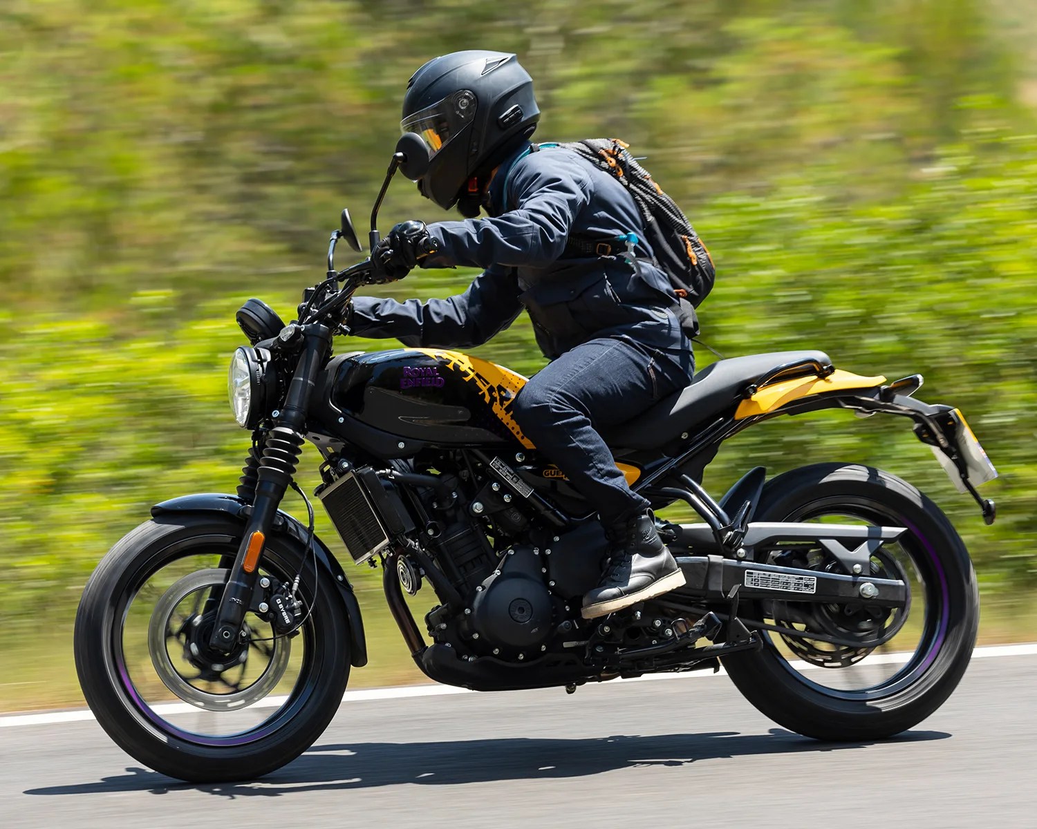 A person wearing a black helmet, dark jacket, jeans, and black boots is riding a black and yellow Royal Enfield motorcycle on a road with a blurred green background. The rider has a black and orange backpack. The motorcycle has a round headlight and visible engine components.