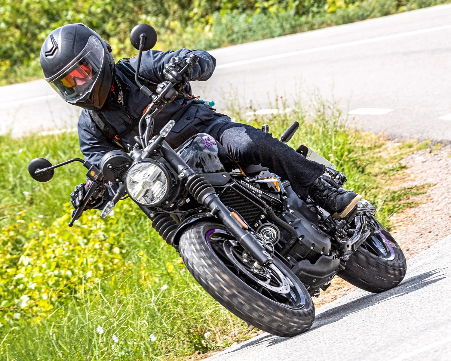 A person wearing a black helmet with a tinted visor, black jacket, gloves, and dark jeans is riding a black motorcycle leaning into a turn on a paved road. The motorcycle has a round headlight and knobby tires, and the background shows green grass and some wildflowers.