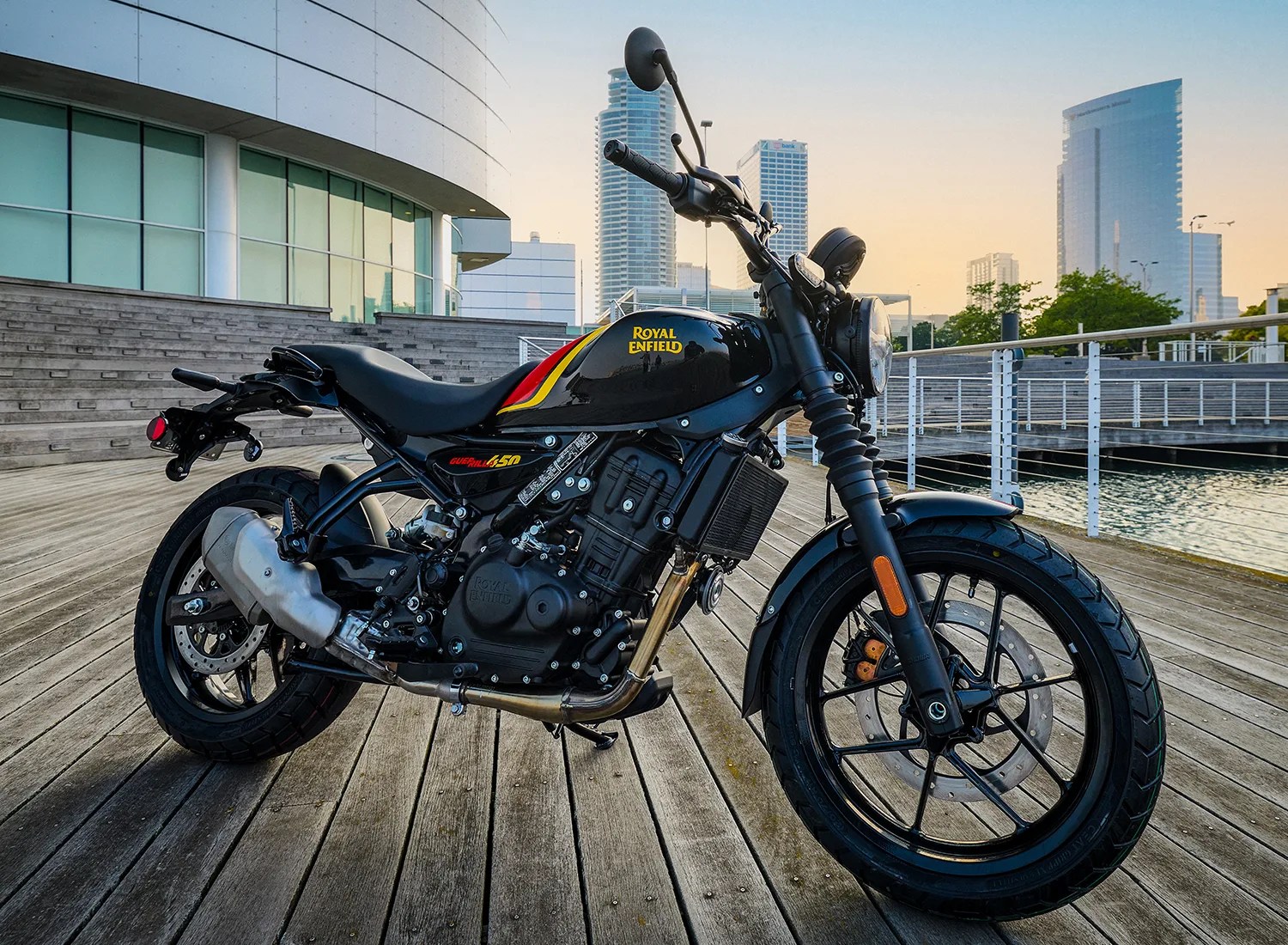 Black Royal Enfield motorcycle with red and yellow stripes on the fuel tank, parked on a wooden deck near a waterfront with modern buildings in the background during sunset.