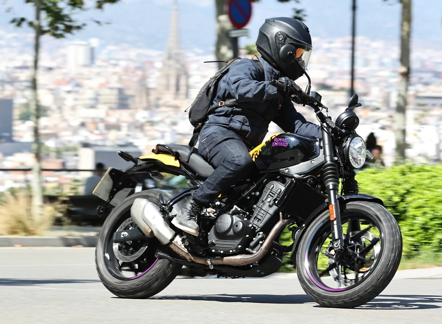 A person wearing a black helmet, dark jacket, jeans, and boots is riding a black and yellow Royal Enfield motorcycle on a city street. The motorcycle has a round headlight and visible exhaust pipes. The background shows a blurred urban landscape with trees and buildings.