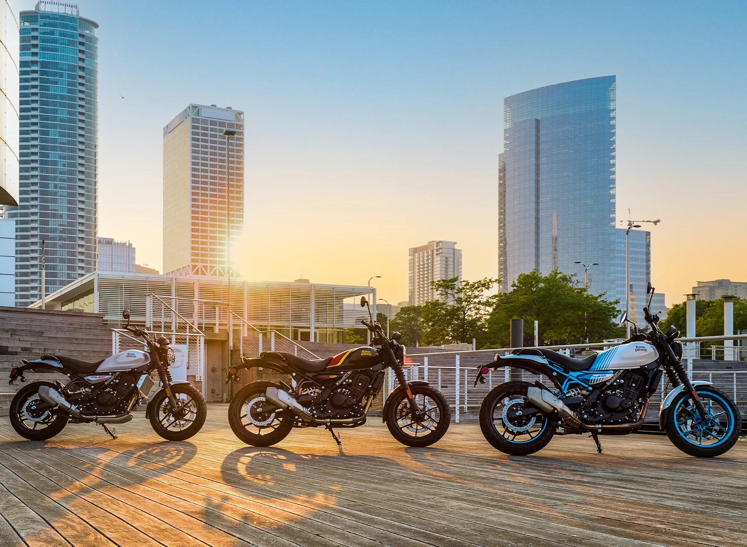 Three Royal Enfield motorcycles are parked on a wooden deck with a city skyline in the background during sunset. The motorcycles have different color schemes: the left one is white with black accents, the middle one is black with yellow and red stripes, and the right one is white with blue accents. The scene is illuminated by the warm light of the setting sun, casting long shadows on the deck. Tall modern buildings and greenery are visible in the background.