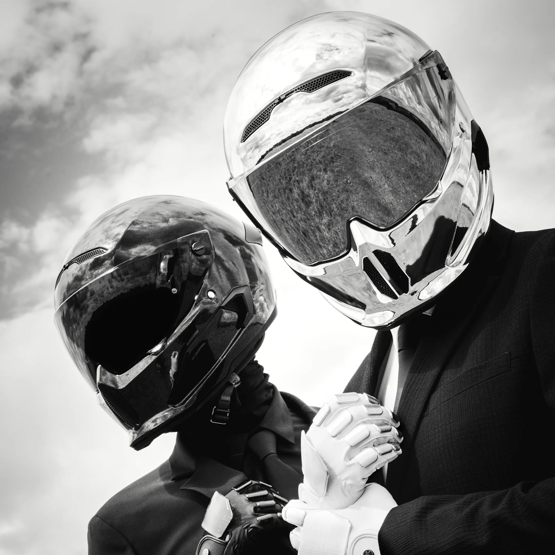 Two individuals wearing reflective motorcycle helmets and black suits with ties are clasping hands. One helmet is shiny silver, and the other is glossy black. Both are wearing gloves matching their helmets, with the person in the silver helmet wearing white gloves and the person in the black helmet wearing black gloves. The background shows a cloudy sky. The image is in black and white.