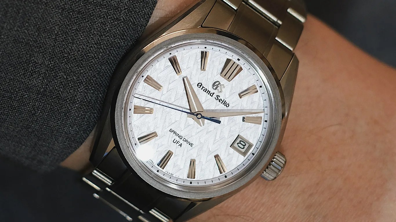 Grand Seiko wristwatch with a textured white dial, silver hour markers, and silver hands. The watch features a date window at 3 o'clock, a blue second hand, and the text "Grand Seiko," "SPRING DRIVE," and "UFA" on the dial. The watch has a titanium case and bracelet.