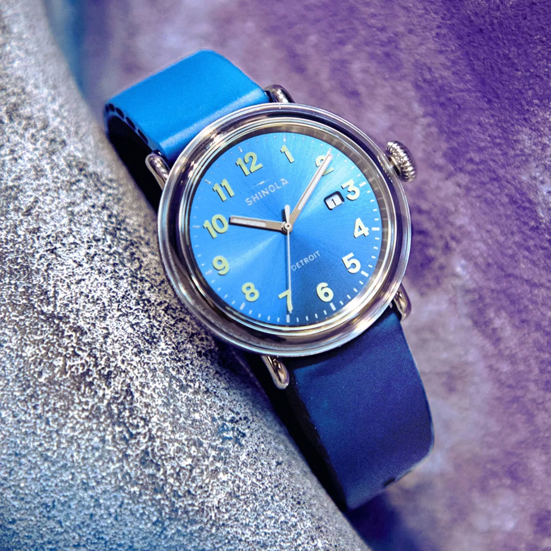 Blue Shinola wristwatch with a round silver case, blue dial, and yellow numerals. The watch has a blue leather strap and features a date window at the 3 o'clock position. The dial also displays the words "SHINOLA" and "DETROIT." The watch hands are silver with a second hand in a lighter shade.