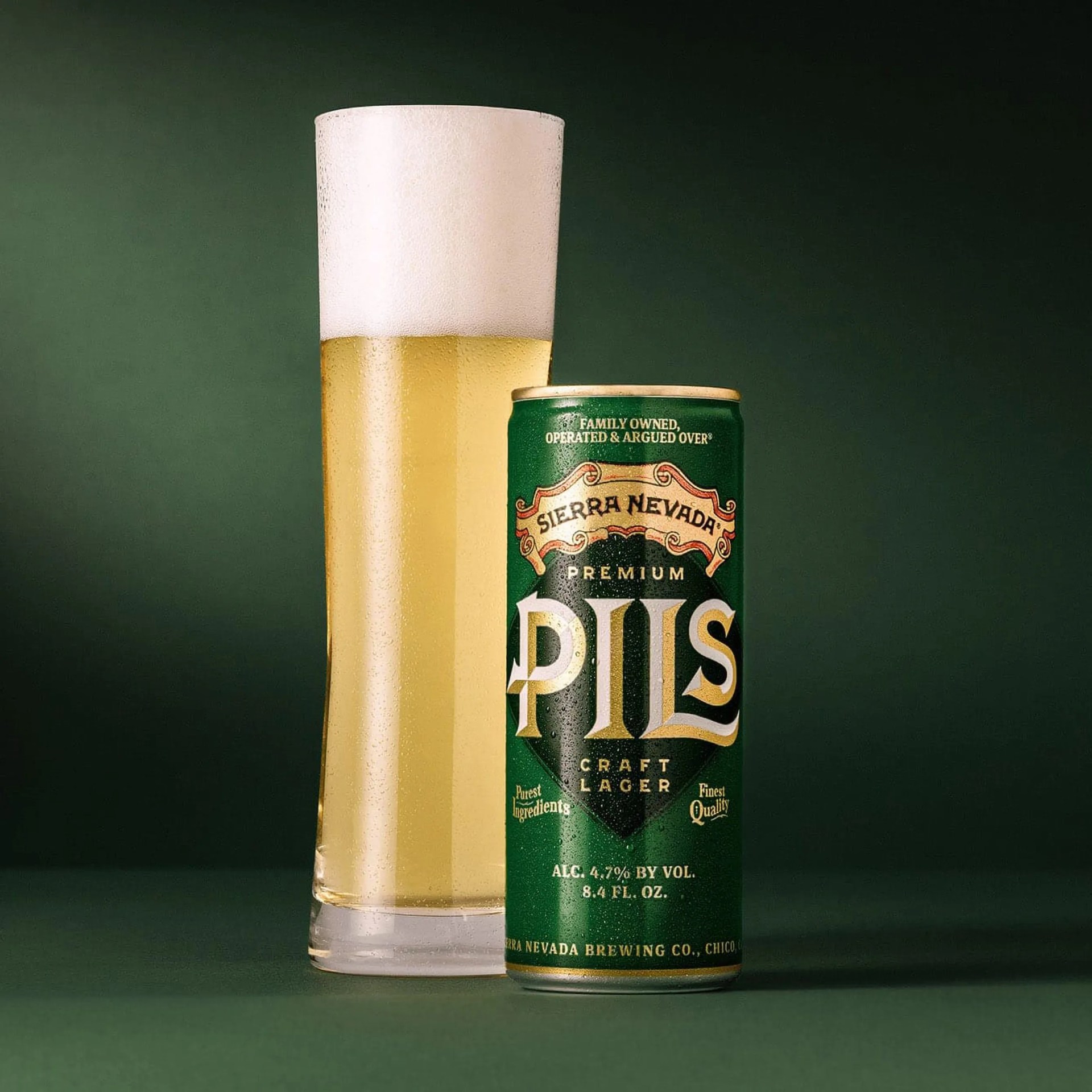 A tall glass of pale golden beer with a thick foamy head next to a green can of Sierra Nevada Premium Pils craft lager. The can features gold and white lettering and states it has 4.7% alcohol by volume. Both the glass and can have condensation droplets.
