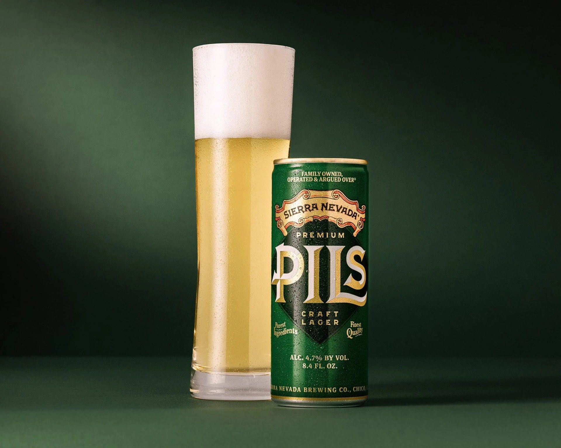 A tall glass of light golden beer with a thick white foam head next to a green can of Sierra Nevada Premium Pils craft lager. The can features gold and black lettering and states it has 4.7% alcohol by volume and contains 8.4 fluid ounces. The background is a dark green color.
