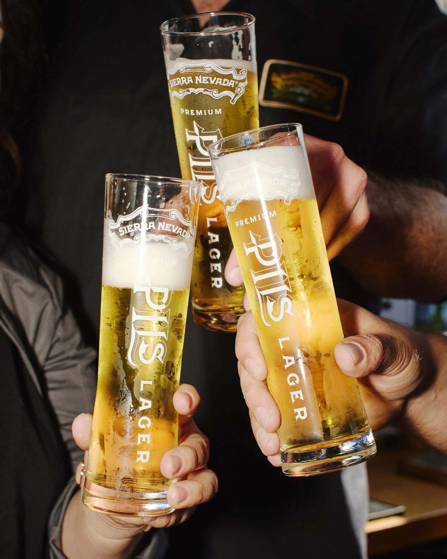 Three hands holding tall, narrow glasses of Sierra Nevada Premium Pils Lager beer, with a light golden color and a foamy head, raised together in a toast.