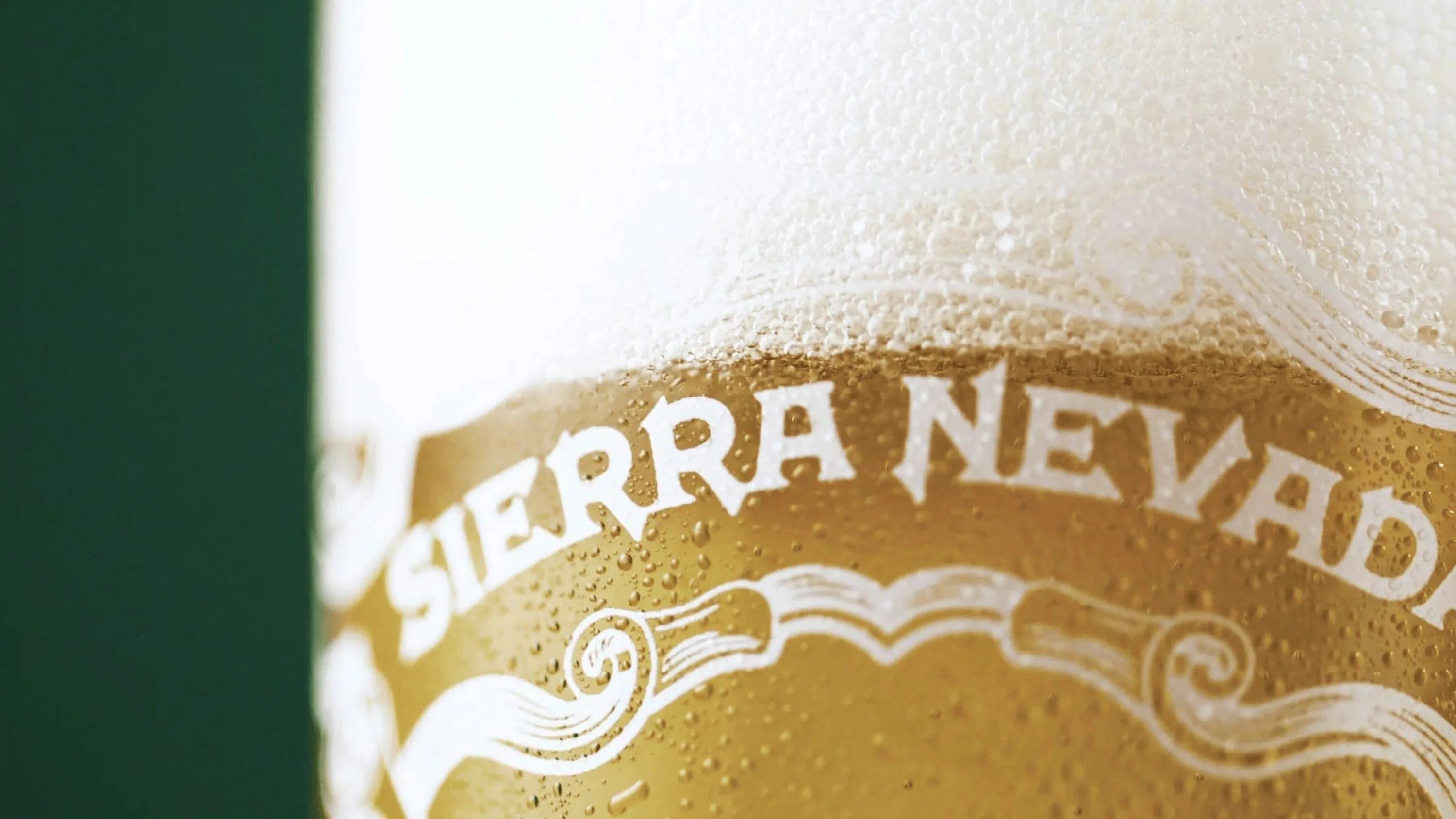 Close-up of a cold glass of Sierra Nevada beer with condensation droplets on the glass and a thick, foamy head on top. The text "SIERRA NEVADA" is prominently visible on the glass.