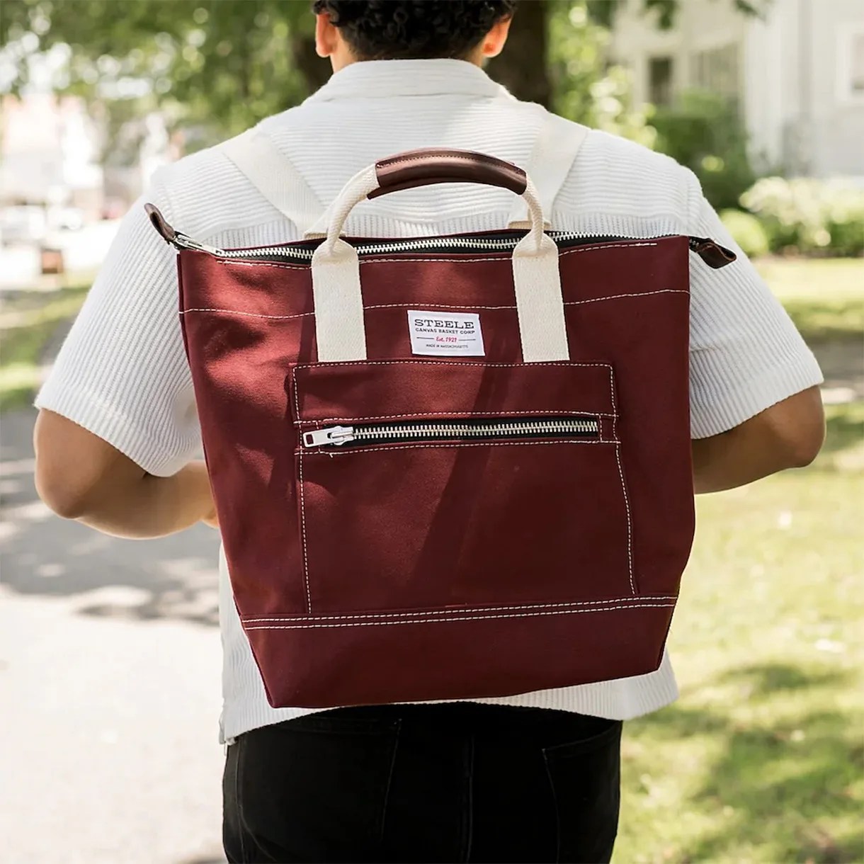 A person wearing a white short-sleeve shirt and black pants carries a maroon canvas backpack with cream-colored handles and a front zippered pocket. The backpack has a label that reads "STEELE CANVAS BASKET CORP." and features white stitching and a leather handle grip.