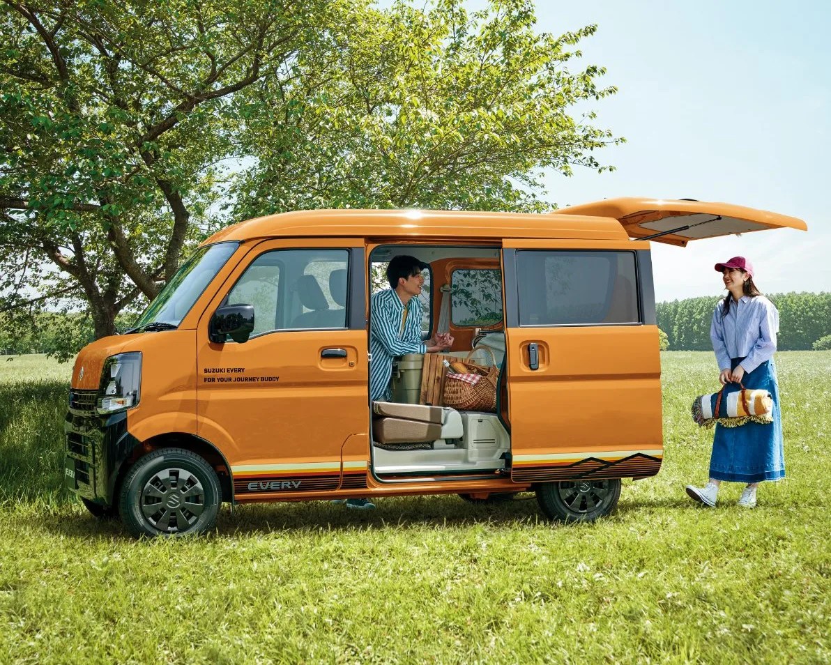 Orange Suzuki Every van parked on grass with its side sliding door and rear hatch open. A man is sitting inside the van near the open sliding door, and a woman wearing a red cap, blue skirt, and striped shirt is standing outside holding a rolled-up blanket. The van is loaded with picnic items including a basket and a cooler. Trees and a clear sky are in the background.