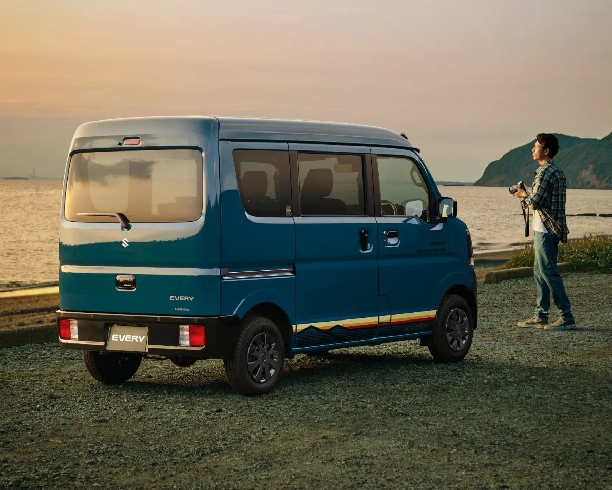 Blue Suzuki Every van parked on a gravel surface near a body of water at sunset, with a man standing nearby holding a camera and looking towards the horizon.