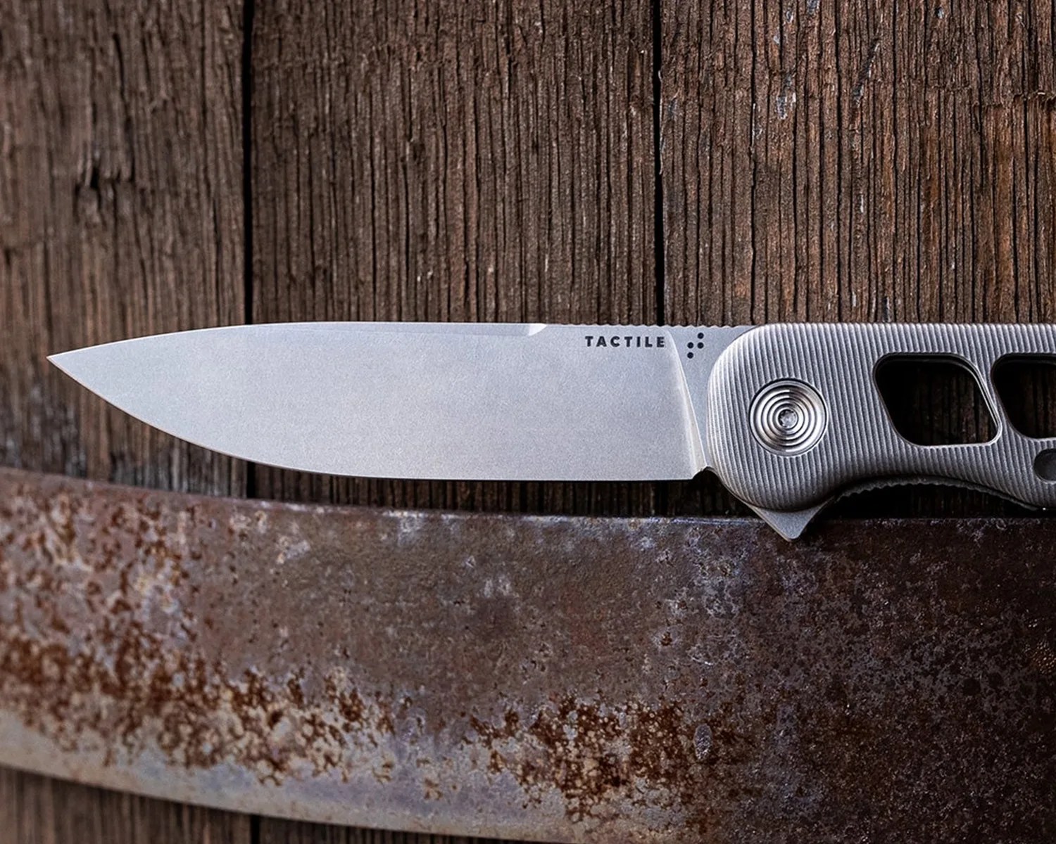 A sleek folding knife with a silver blade and a textured silver handle. The blade has the word "TACTILE" printed near the handle. The handle features circular grooves around the pivot and cut-out sections for grip. The knife is resting on a rustic wooden surface with a rusty metal edge below it.