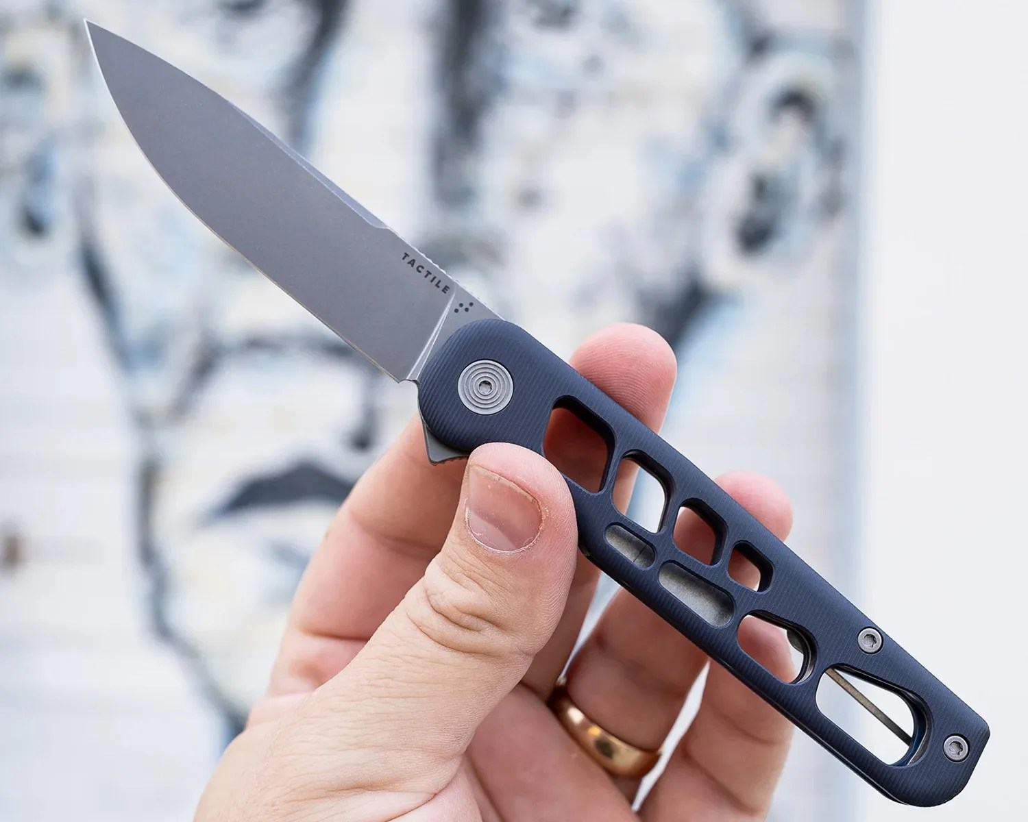 A hand holding a folding knife with a matte silver blade and a black skeletonized handle. The blade has the word "TACTILE" printed near the base. The handle features multiple cutouts and a circular pivot with concentric rings. The person holding the knife is wearing a gold ring on their ring finger.