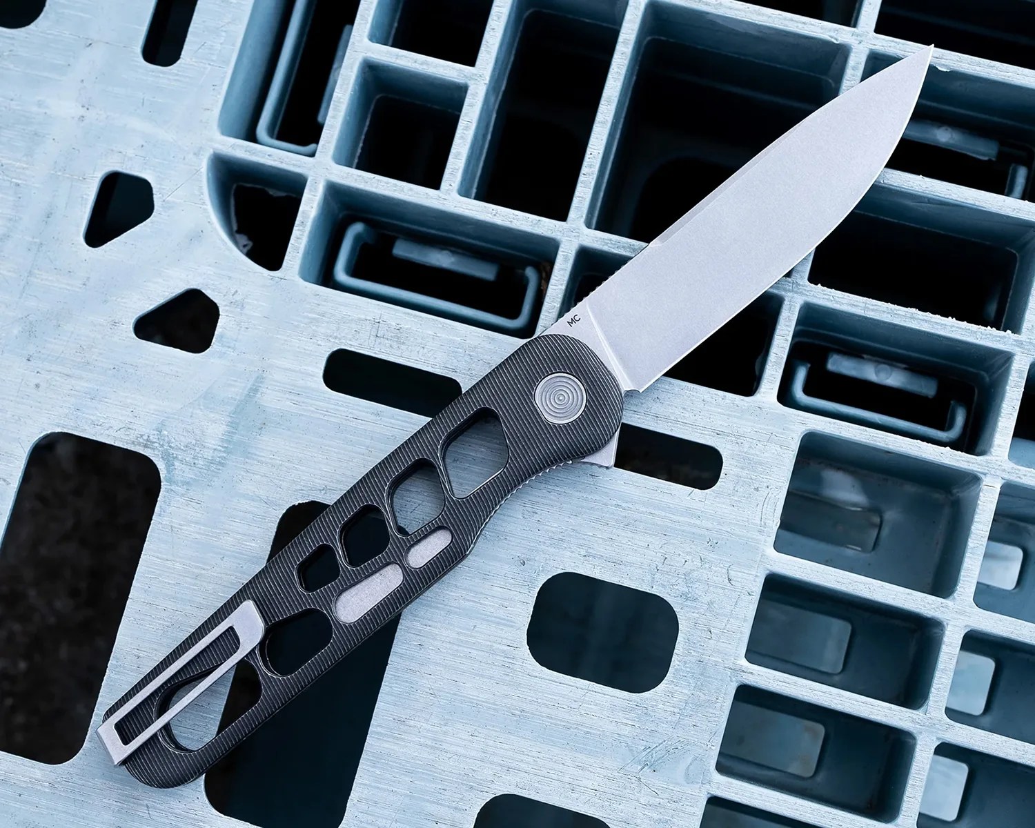 Folding knife with a sleek, modern design featuring a silver blade and a black handle with multiple cut-out sections and textured grip, resting on a light blue plastic grid surface.