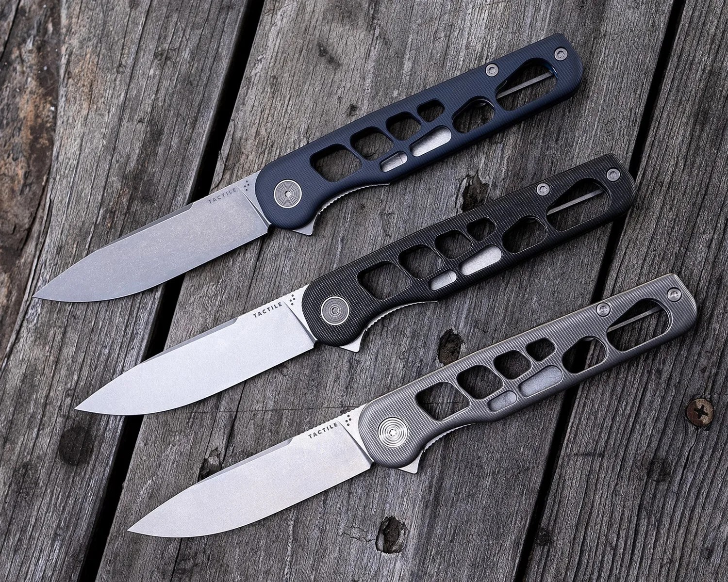 Three folding knives with stonewashed blades and skeletonized handles in different shades of black and gray, arranged diagonally on a weathered wooden surface. The blades are marked with the brand name "TACTILE.