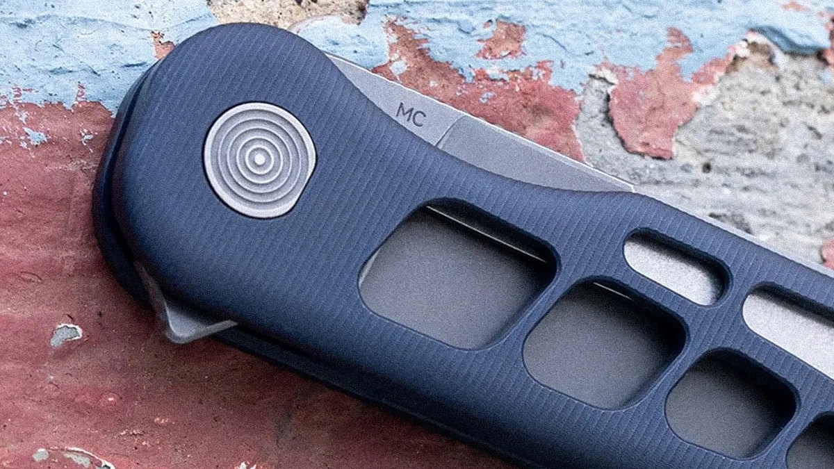 Close-up of a folding knife handle with a textured dark blue grip, featuring a circular metal pivot with concentric rings and cut-out sections along the handle. The blade is partially visible, marked with "MC." The background is a weathered surface with peeling paint in blue, red, and beige tones.