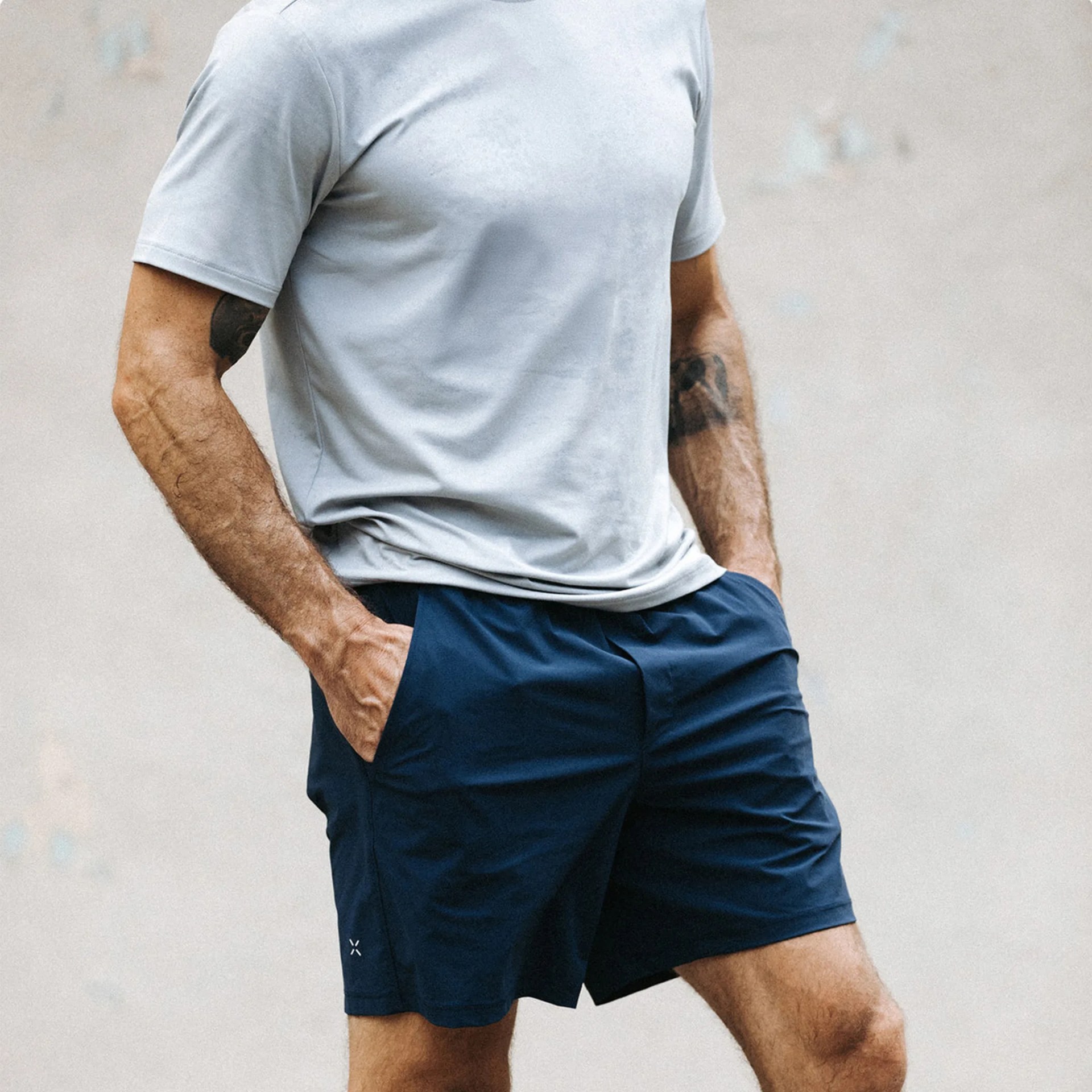 A man wearing a light gray short-sleeve t-shirt and navy blue athletic shorts with his hands in the shorts' pockets. The shorts have a small white logo near the bottom hem on the left leg. The background is plain and out of focus.
