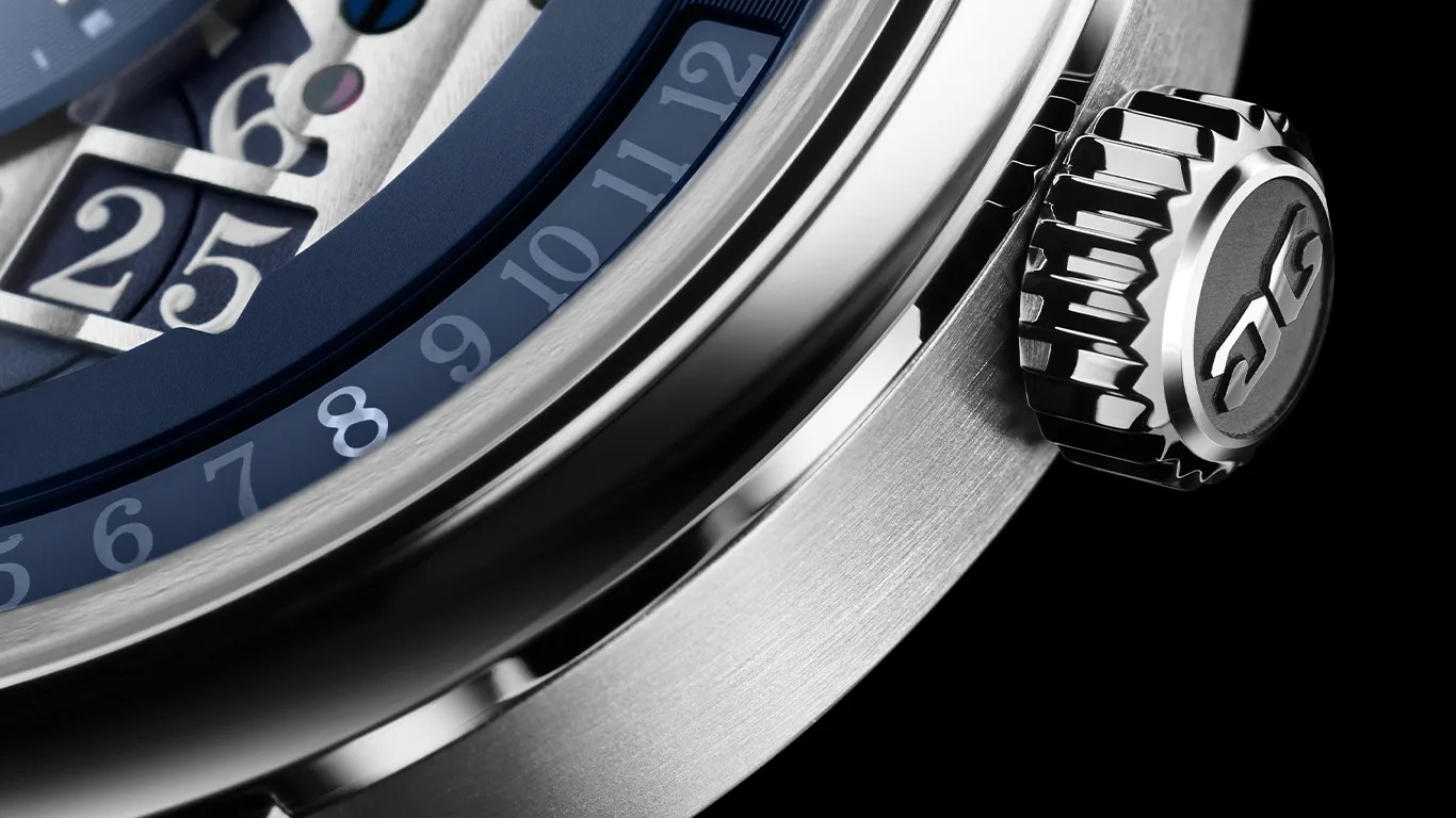 Close-up of a glashutte original watch crown and part of the bezel, showing a brushed metal finish and a blue ring with white numerals from 6 to 12. The crown features a textured edge and an embossed logo. The watch face includes a date window displaying the number 25.