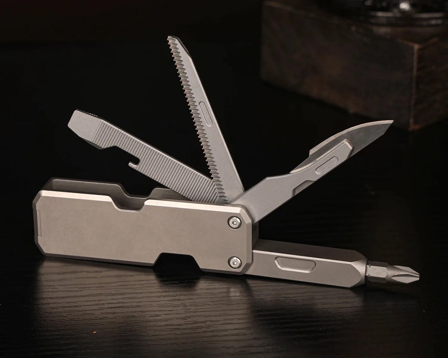 Compact multi-tool with a metallic finish, featuring an open knife blade, serrated saw, flathead screwdriver, and a Phillips screwdriver bit, all extended from a rectangular handle. The tool is placed on a dark wooden surface with a blurred background.