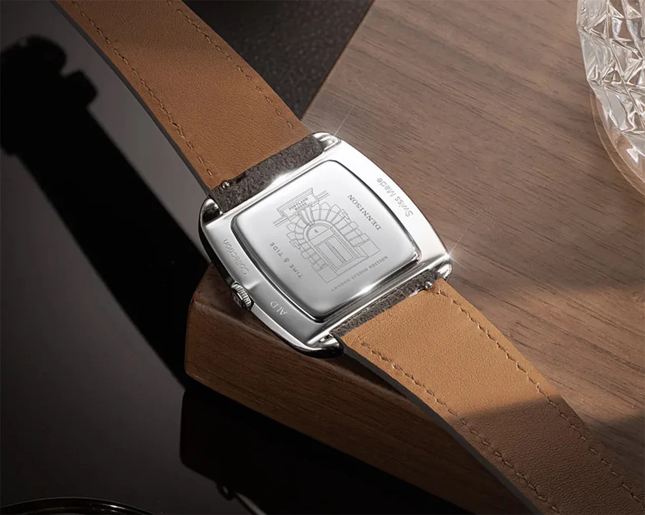 A close-up of the back of a wristwatch with a polished silver case and a tan leather strap. The case back features an engraved architectural design and text including "DENNISON," "TIME & TIDE," "SWISS MADE," "AID," and "Collection." The watch is resting on a wooden surface with a glass object partially visible on the right.