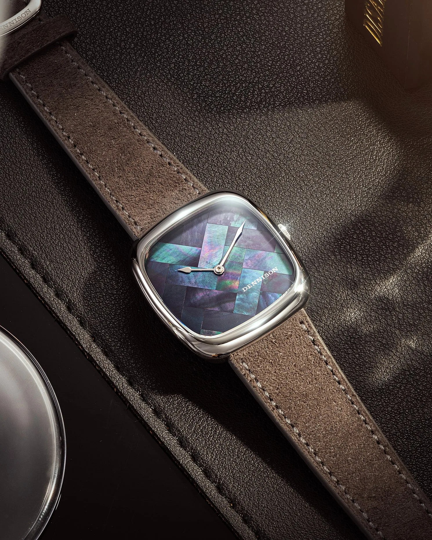 Square wristwatch with a polished silver case and a unique iridescent mosaic dial in shades of blue and purple. The watch has silver hour and minute hands and the brand name "DENNISON" printed on the dial. It features a brown suede leather strap with visible stitching, placed on a textured dark brown leather surface.