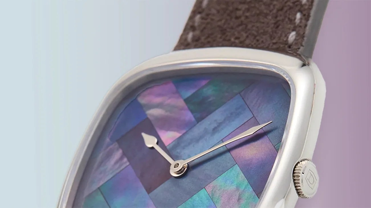 Close-up of a wristwatch with a silver case and a brown leather strap. The watch face features a colorful, iridescent mosaic pattern in shades of blue, purple, and green, with silver hour and minute hands. The crown on the side of the case has a small engraved logo.