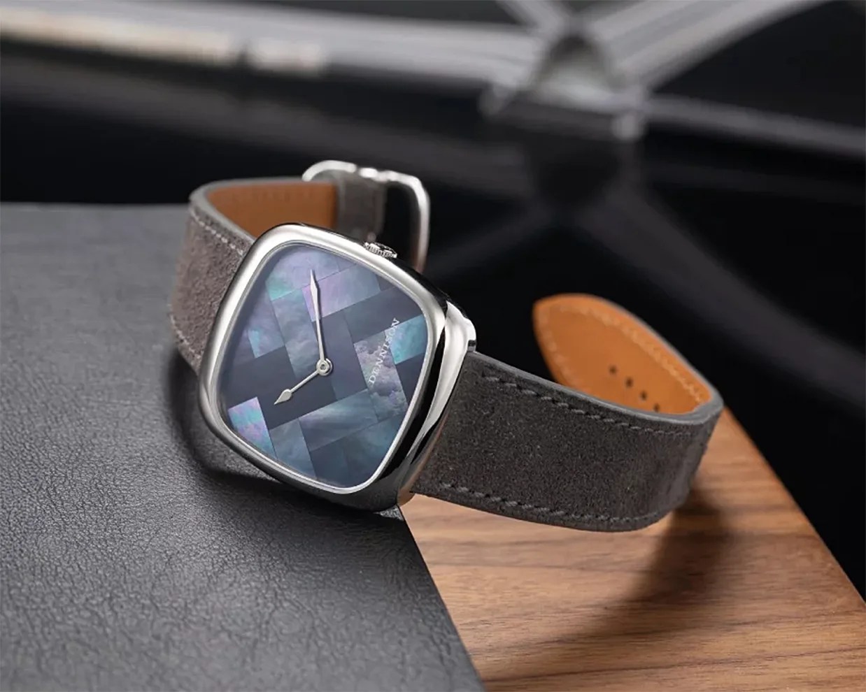 A wristwatch with a square, silver-colored case and a dark, iridescent, mosaic-patterned dial. The watch has silver hands and a gray suede strap with visible stitching and a tan underside. The watch is displayed on a surface combining black leather and wood textures.
