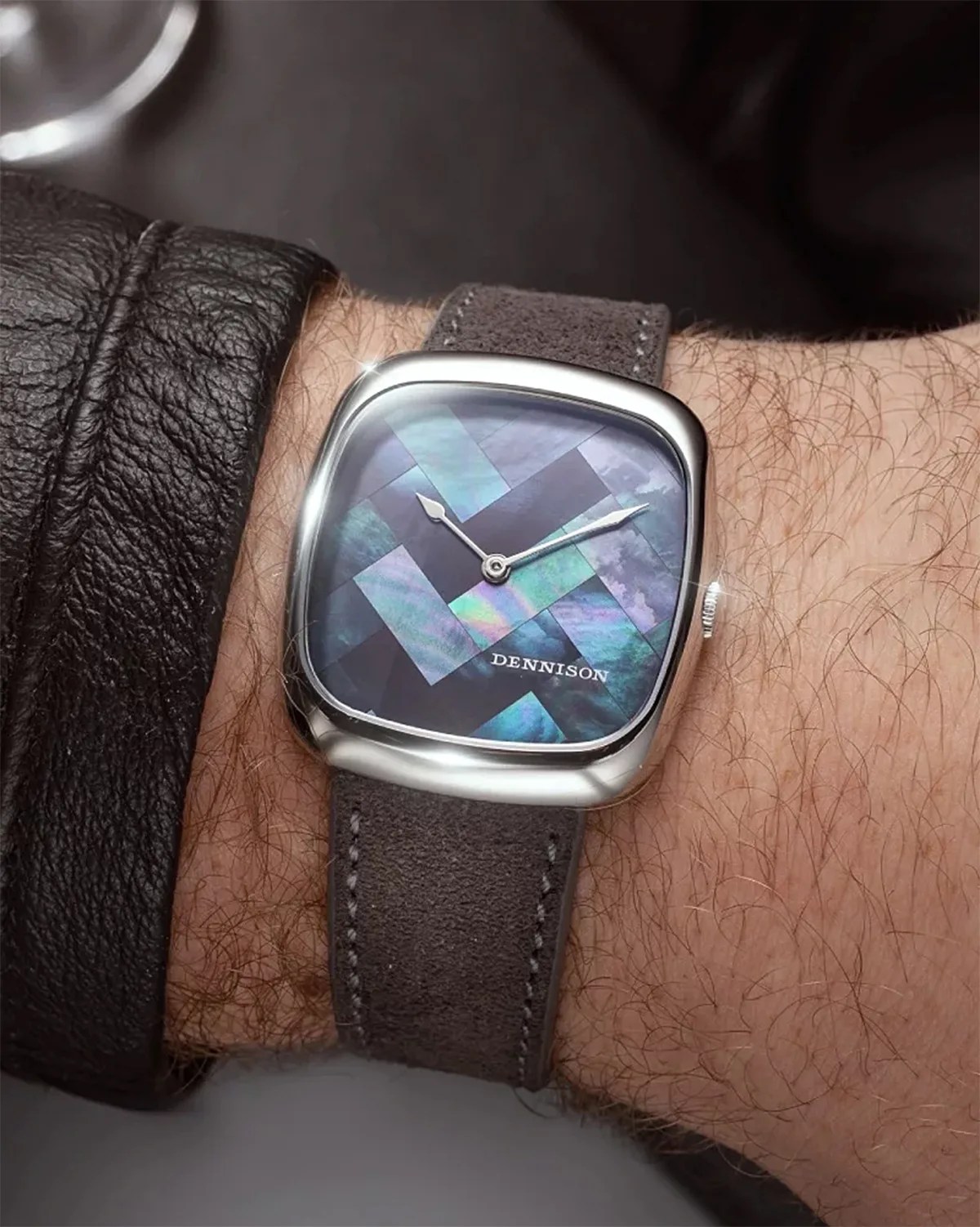 Square wristwatch with a polished silver case and a mosaic-style iridescent blue and green dial. The watch face features silver hour and minute hands and the brand name "DENNISON" printed near the bottom. The strap is dark gray suede with visible stitching. The watch is worn on a wrist with a black leather jacket sleeve partially visible.